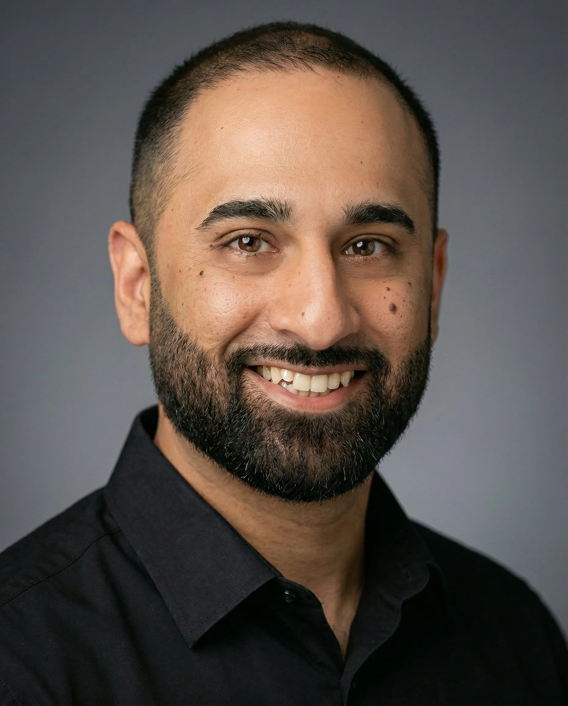 Close-up portrait of a smiling man with a beard, dark hair, and brown eyes, dressed in a black shirt against a gray background.