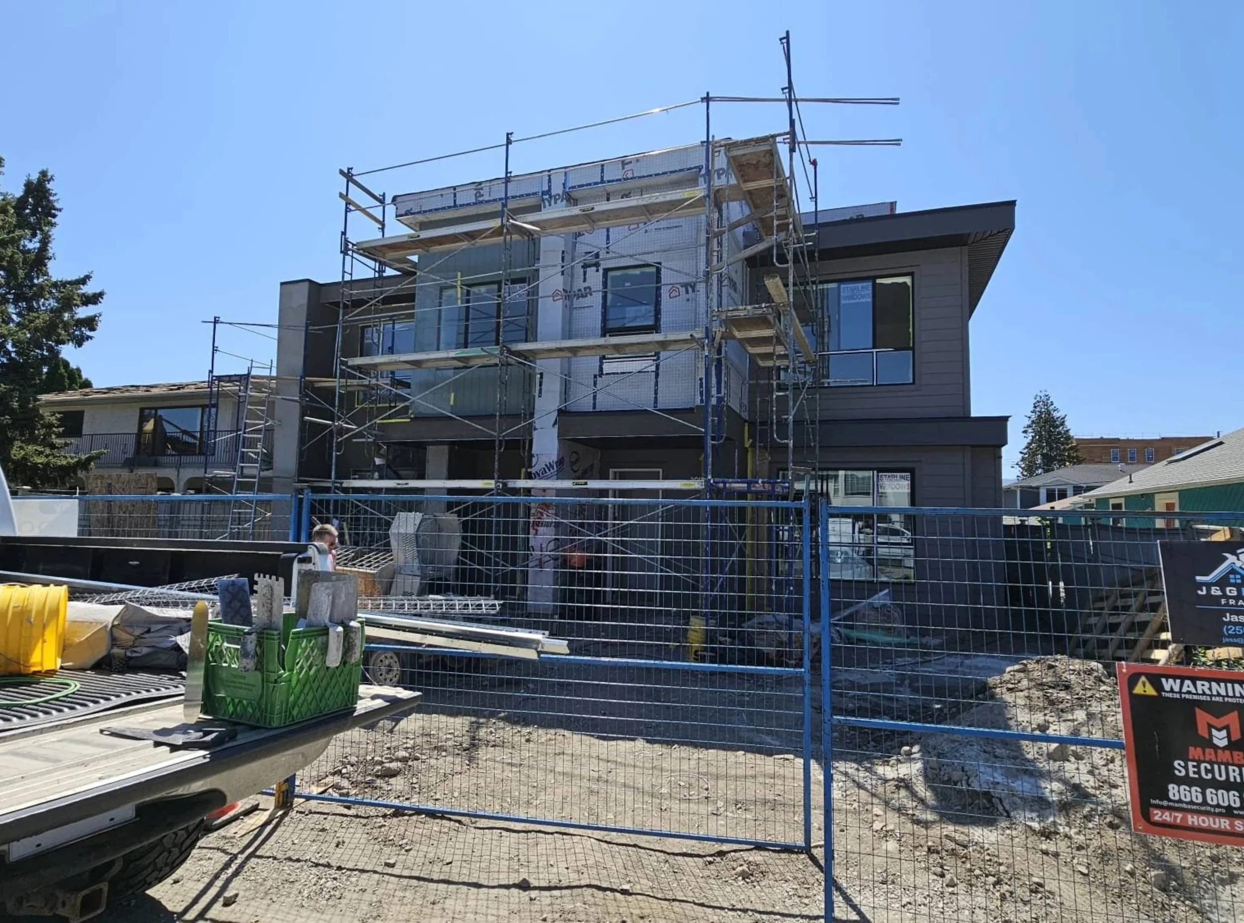 Building under construction with scaffolding around it, partially covered with insulation material, in a residential neighborhood on a sunny day.