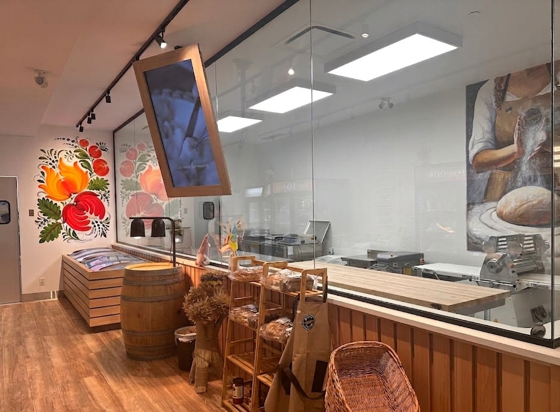 Interior of a bakery or cafe with colorful floral mural, glass wall, and kitchen area. There's a large TV, barrel, and wooden chairs.