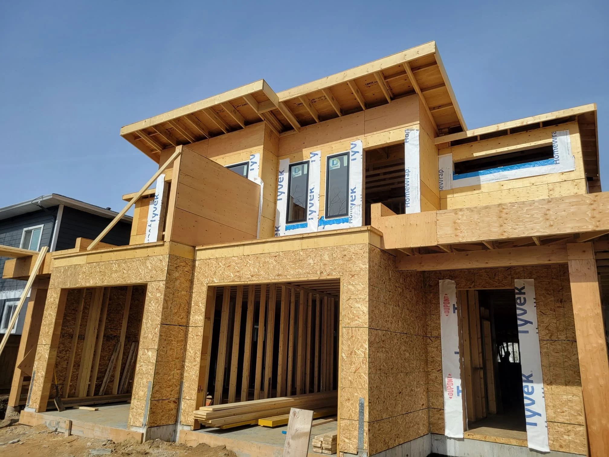 Construction of a multi-story house with wooden framing and scaffolding, in progress, under a clear blue sky.