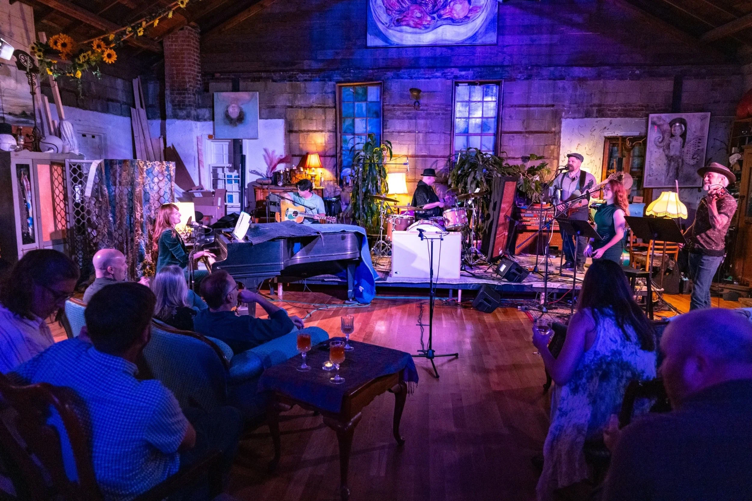 Musicians performing on a stage in a cozy, rustic venue with wooden walls, indoor plants, and warm lighting, while an audience watches and listens.