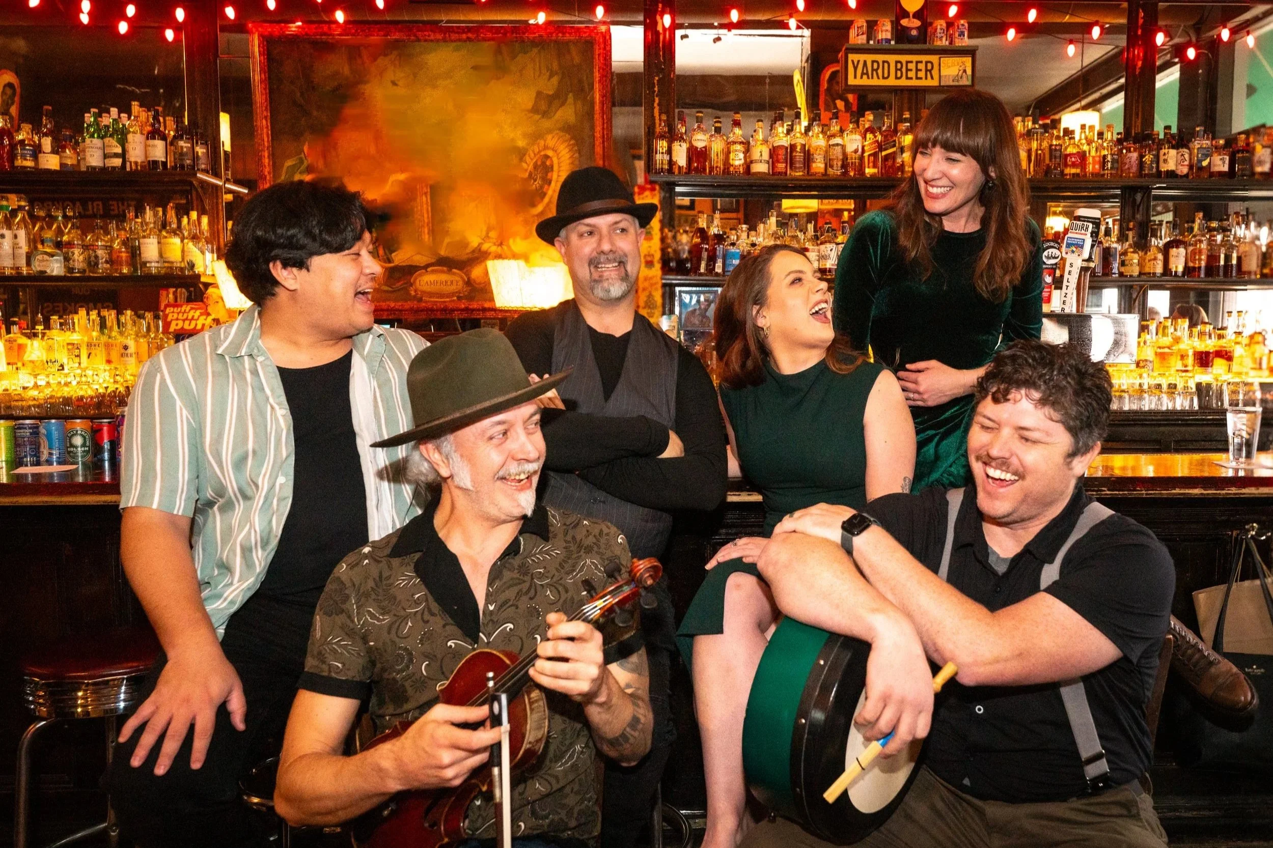 A group of six friends enjoying themselves at a bar, some with musical instruments, laughing and talking with each other, with a well-stocked bar in the background.
