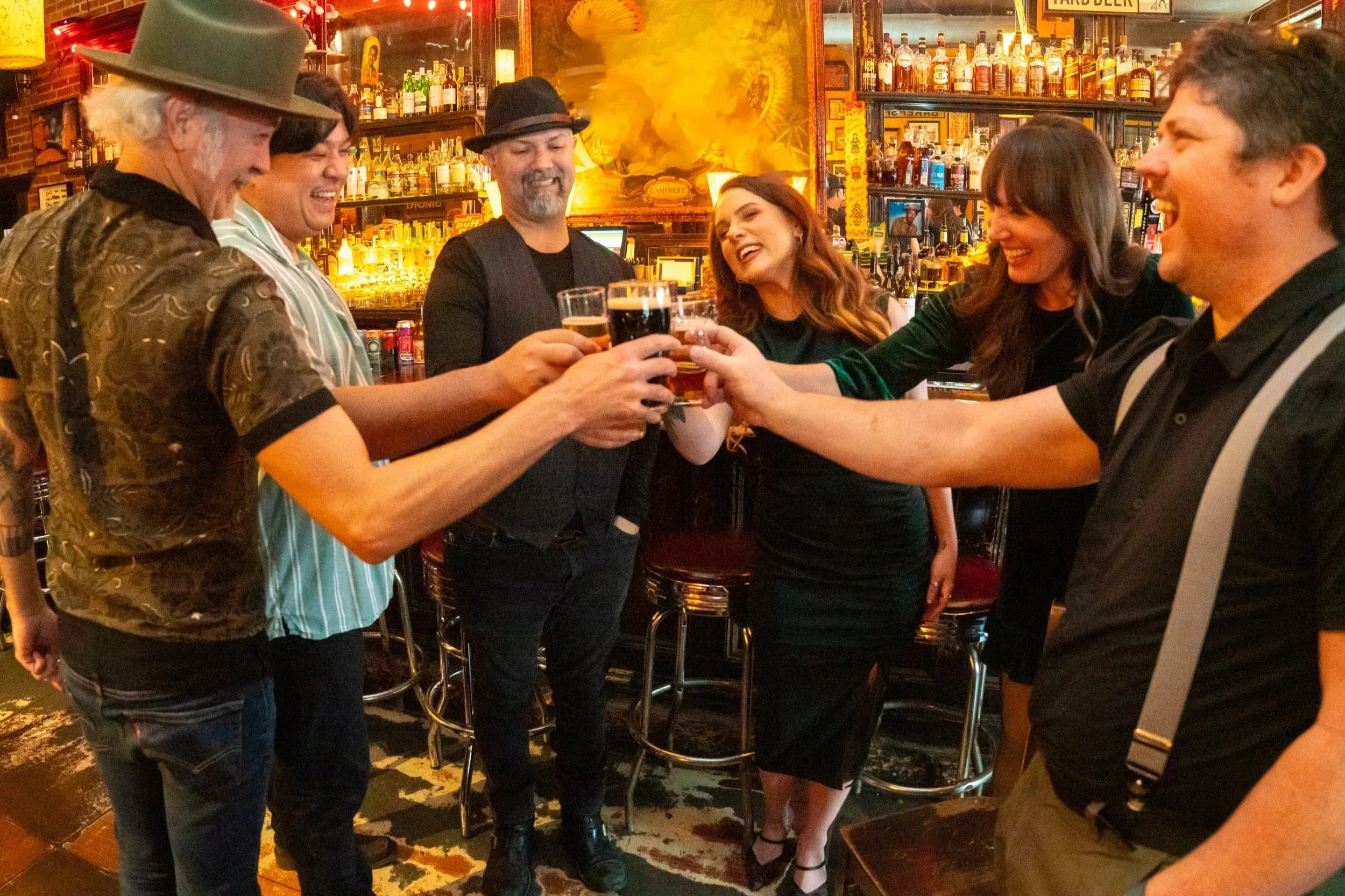 Group of friends toasting drinks in a bar with warm lighting and bottles on shelves in the background.