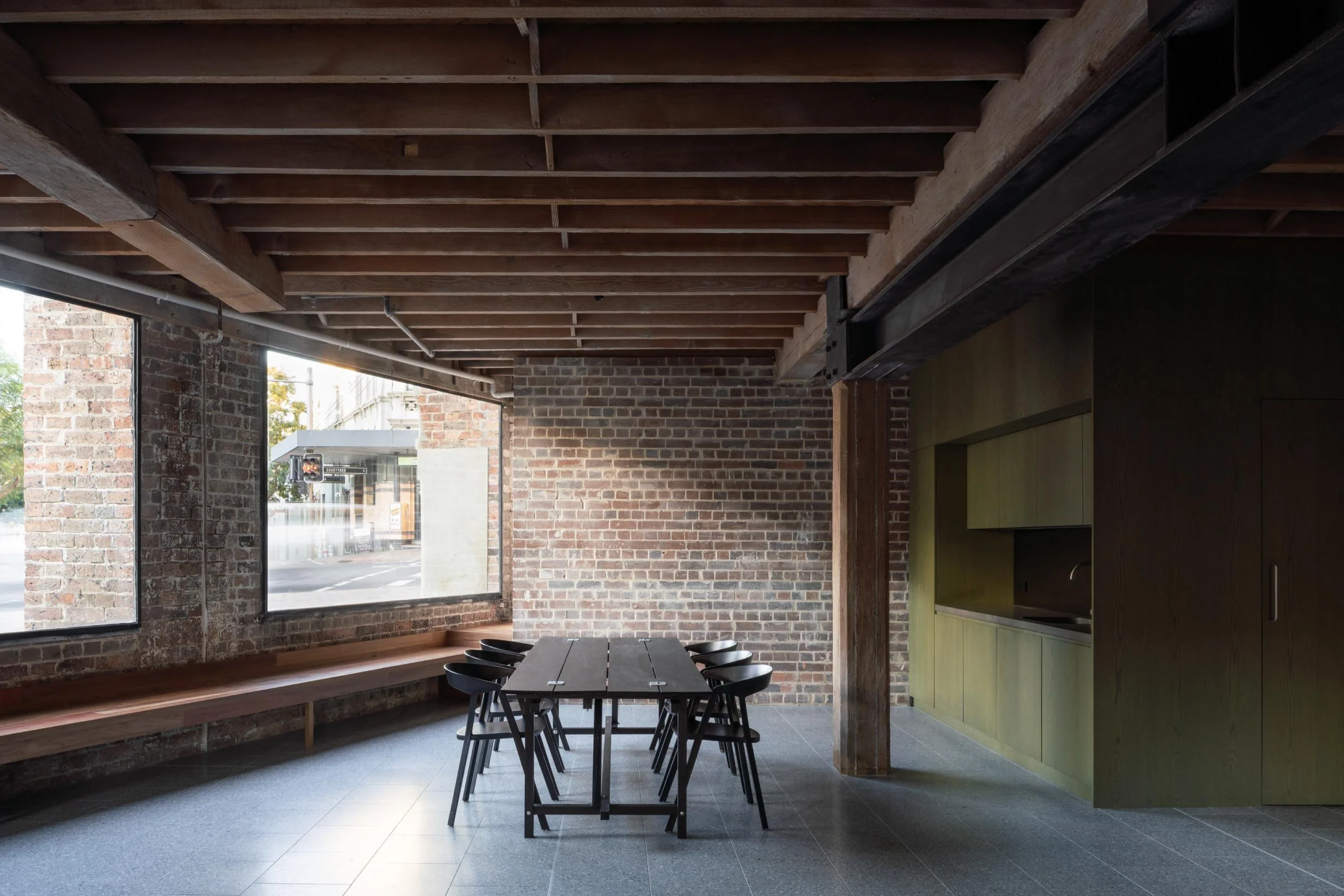 A wide shot of the The Foundry office fitout in Darlington, Sydney, by Muci architects. On the left is a blackbutt bench seat and on the right is a stained green timber joinery kitchen. Fibonacci stone jagger tiles on the floor.
