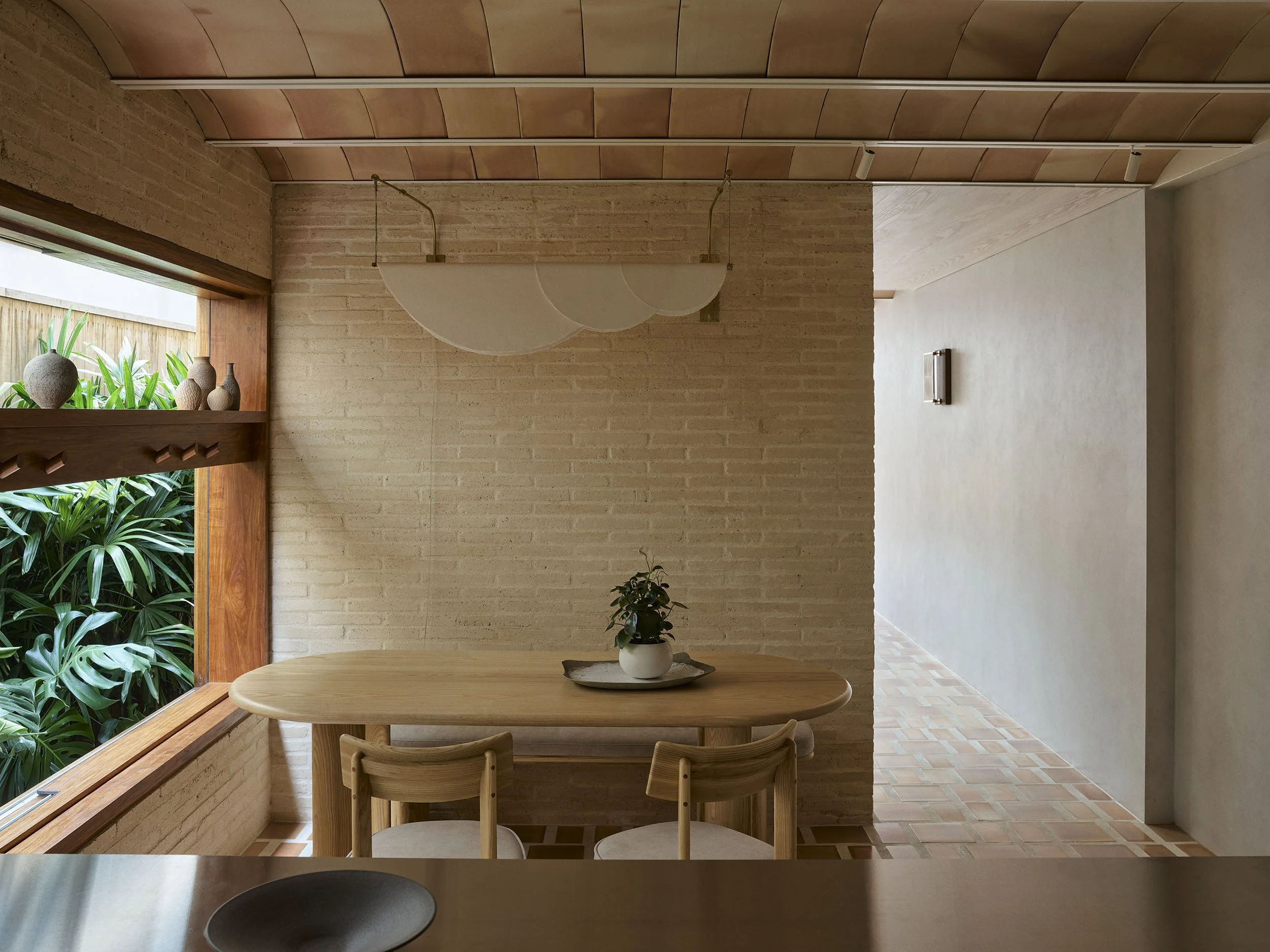 The dining room of Clay House by architect in Newcastle, Muci. A background of cream bricks, a terracotta floor and ceiling, with timber windows and views to gardens.