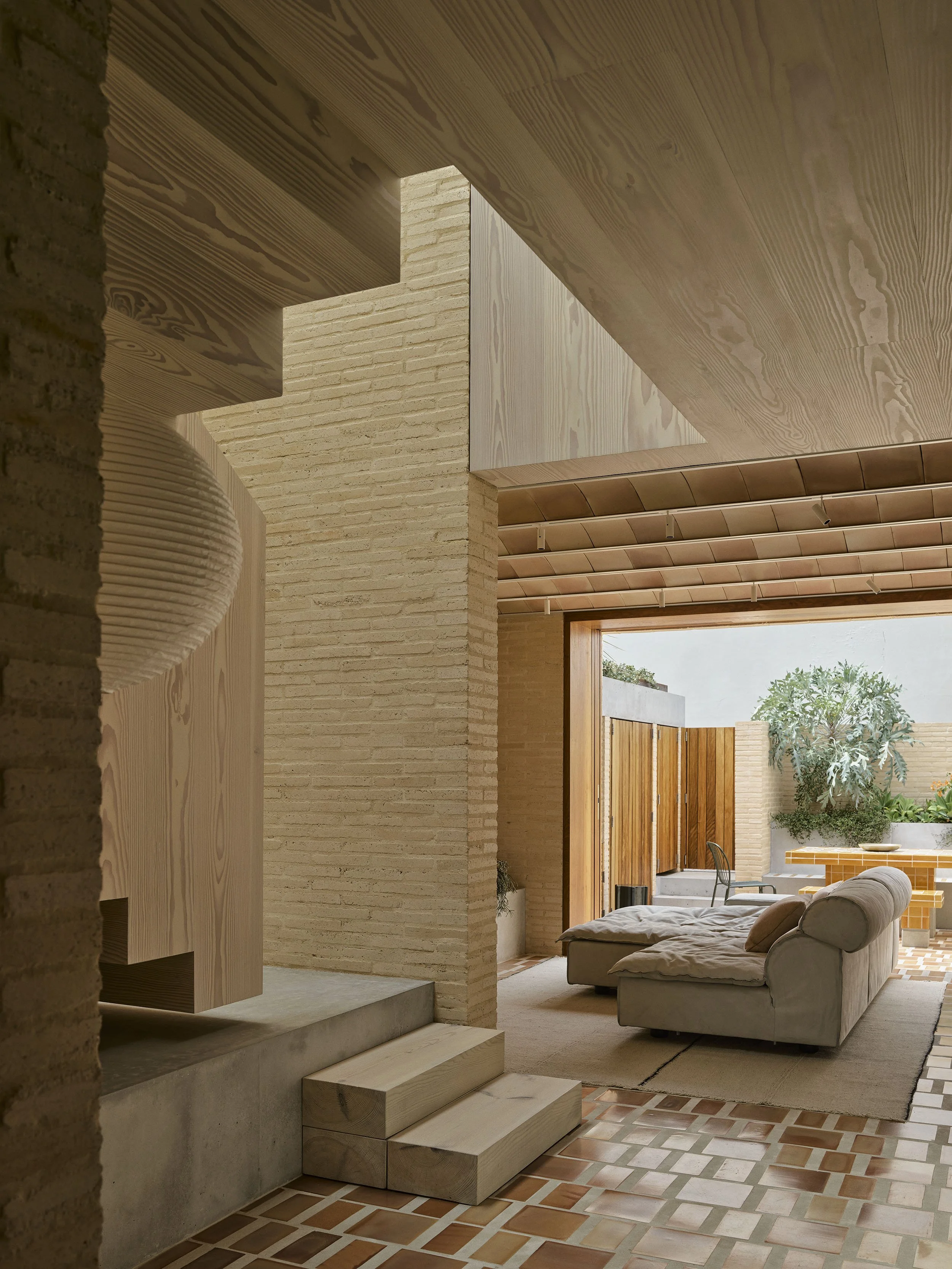 A view at the base of the stairwell in Clay House Sydney terrace renovation by Muci architects. There is a terracotta floor, timber stairs with a zig-zag bottom and cream bricks. Plus a baxter Miami sofa in the living room beyond.