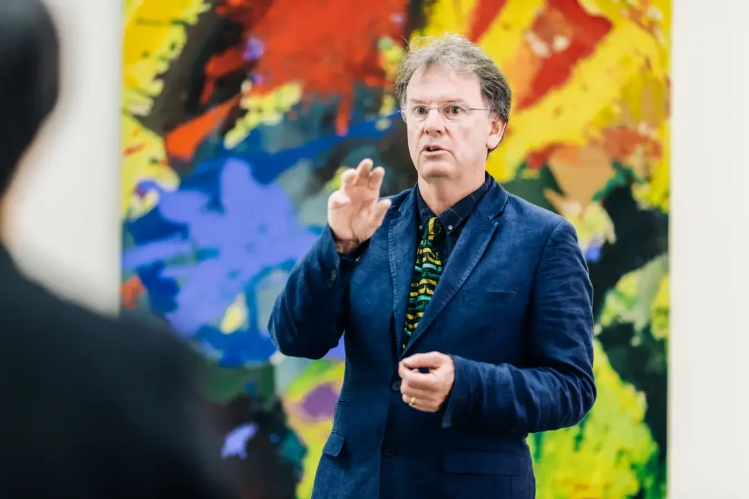 A man with glasses and gray hair wearing a blue blazer, gesturing while speaking, standing in front of an abstract colorful painting.