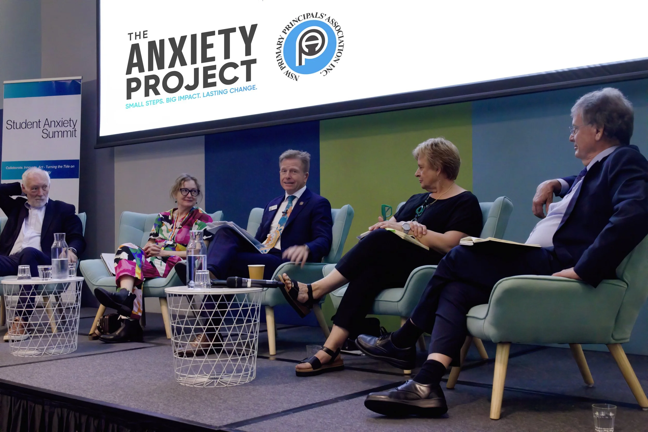 A panel discussion at the Student Anxiety Summit with four people sitting on stage, three women and one man, engaged in conversation. The stage has a large screen with the logo of the Anxiety Project and the text 'Small Steps. Big Impact. Lasting Change.' and a banner reading 'Student Anxiety Summit.' There are small white wire mesh tables and water glasses in front of them.