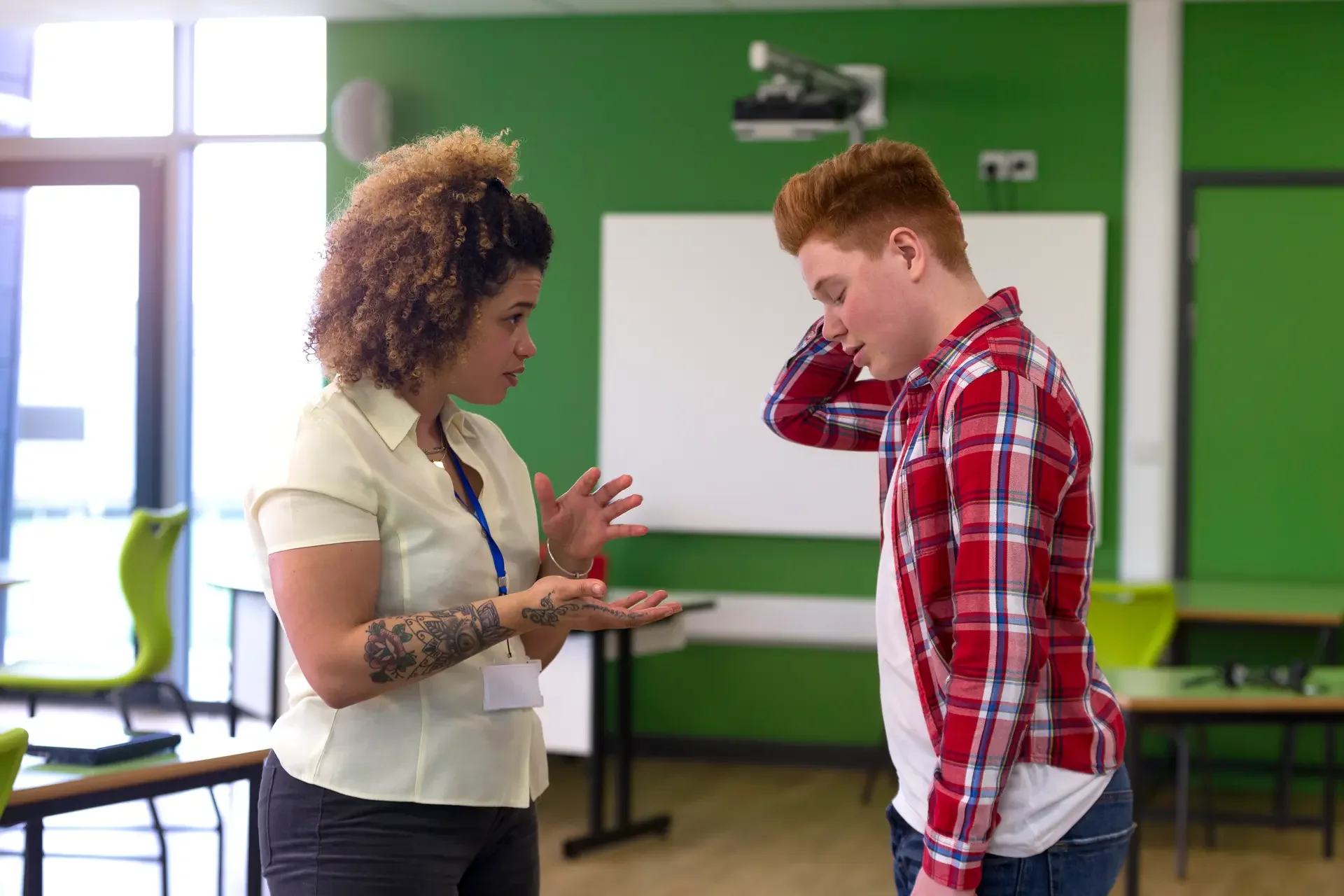 A woman with tattoos on her arm talks to a young man wearing a red and plaid shirt in a classroom or office with green walls and modern chairs.