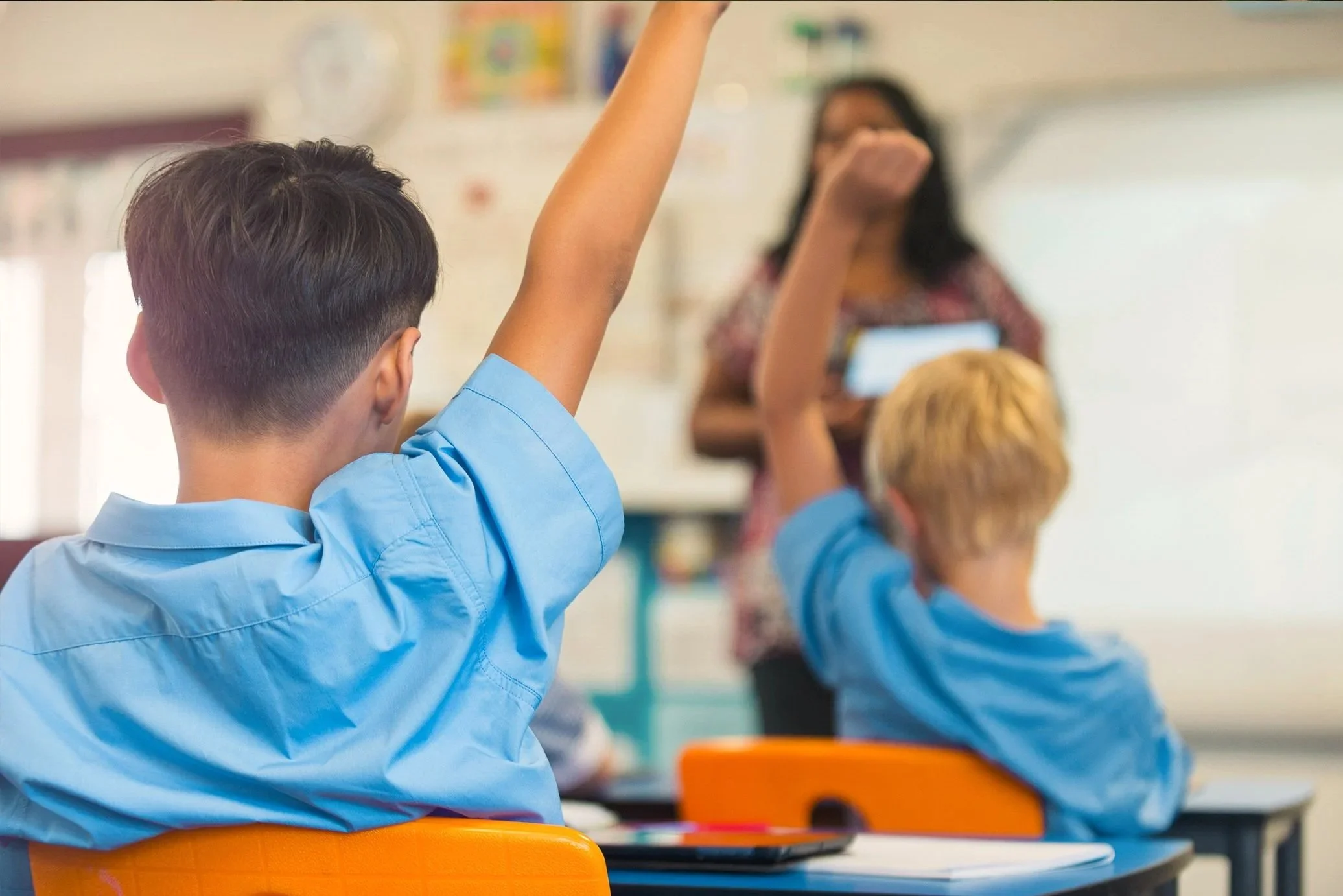 A child in a blue shirt with short dark hair raises his hand in a classroom. In the background, a woman with glasses and a patterned top holds a book and appears to be teaching. The classroom has colorful decorations and furniture.