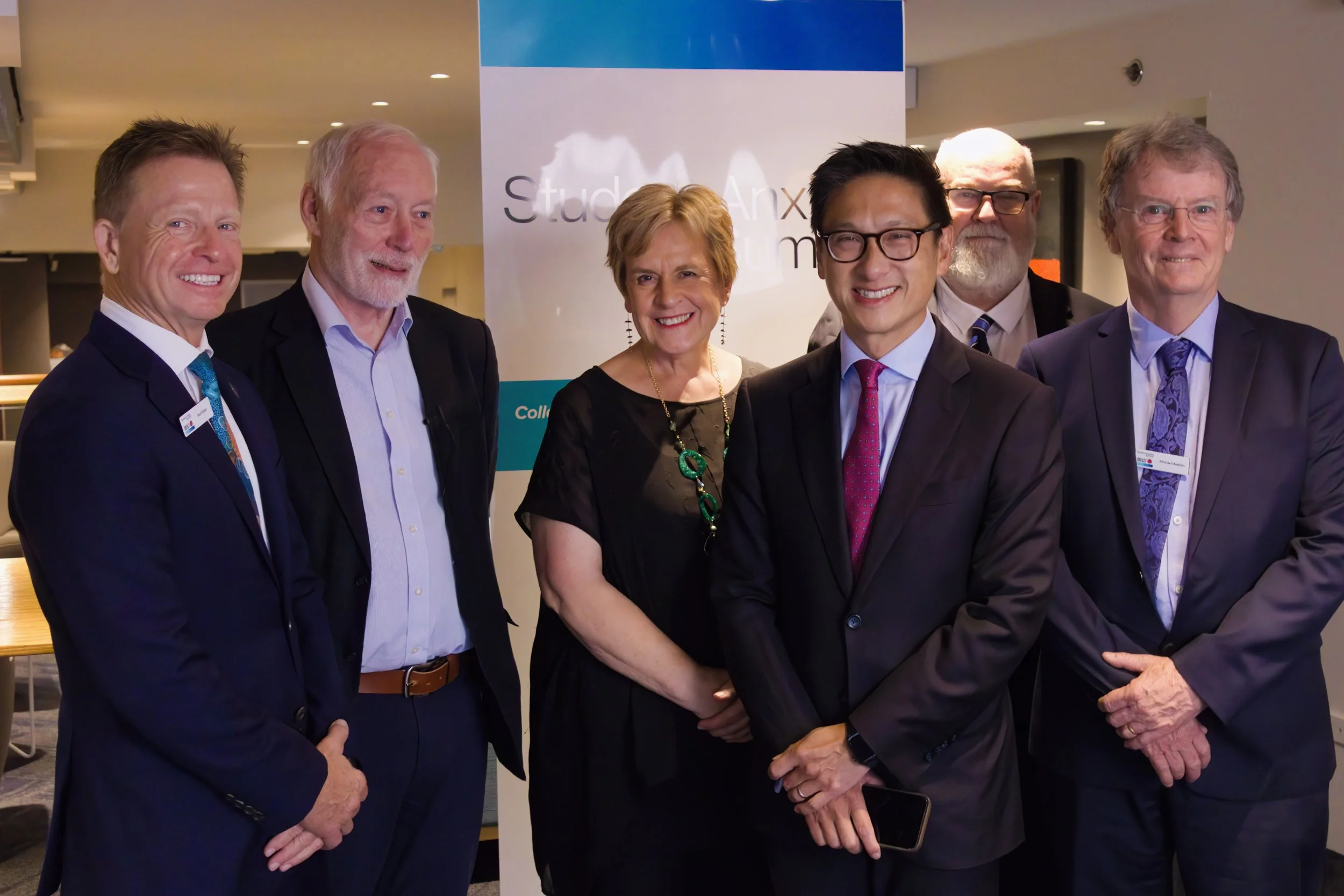 Group of six people in professional attire posing for a photo at a conference with a banner behind them.