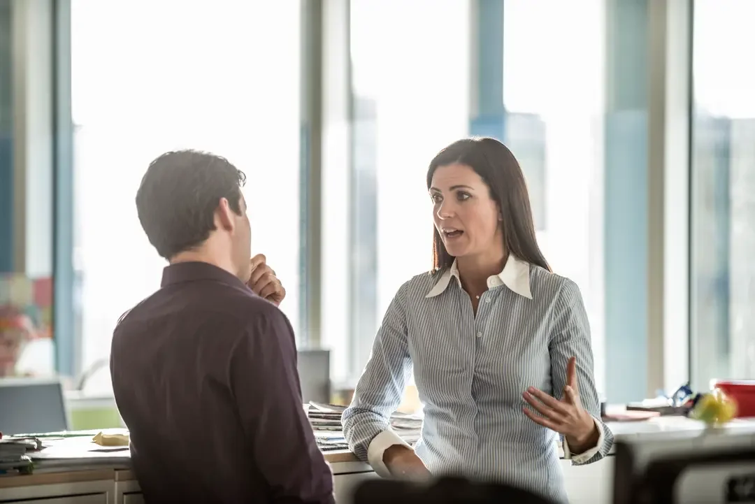 Two people having a conversation in an office, with large windows in the background.