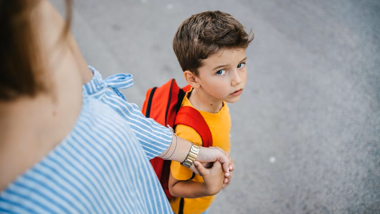 A young boy with brown hair, blue eyes, wearing a yellow shirt and red backpack, looking up at the camera while holding hands with an adult.
