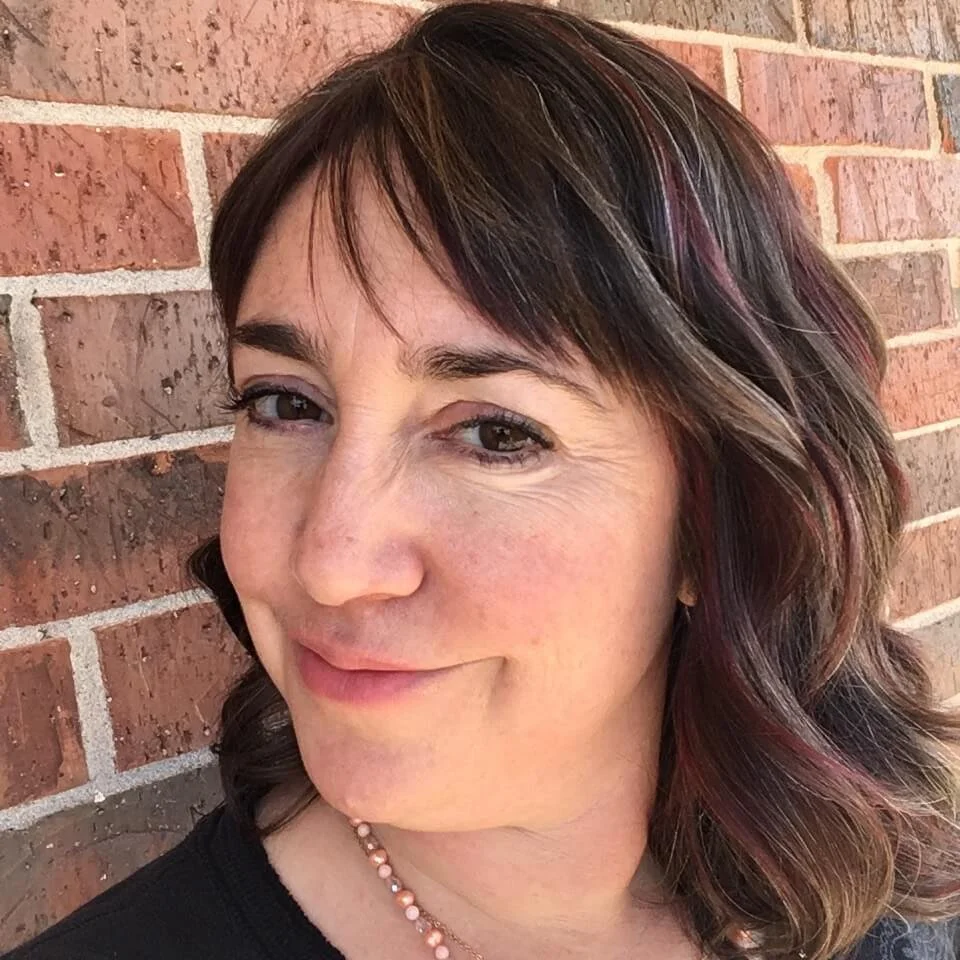 Close-up of a woman with short, wavy brown hair with pink highlights, smiling and leaning against a brick wall.