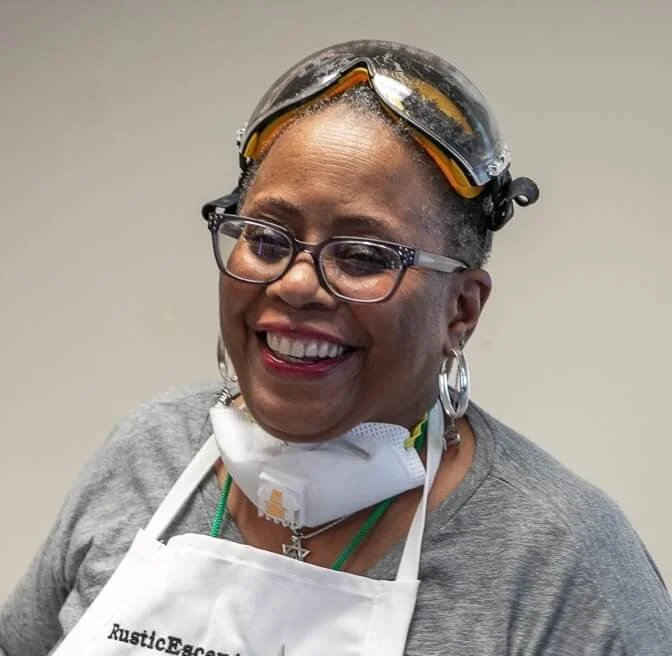 Smiling woman wearing safety glasses, goggles on her head, a face mask around her neck, an apron, and hoop earrings.