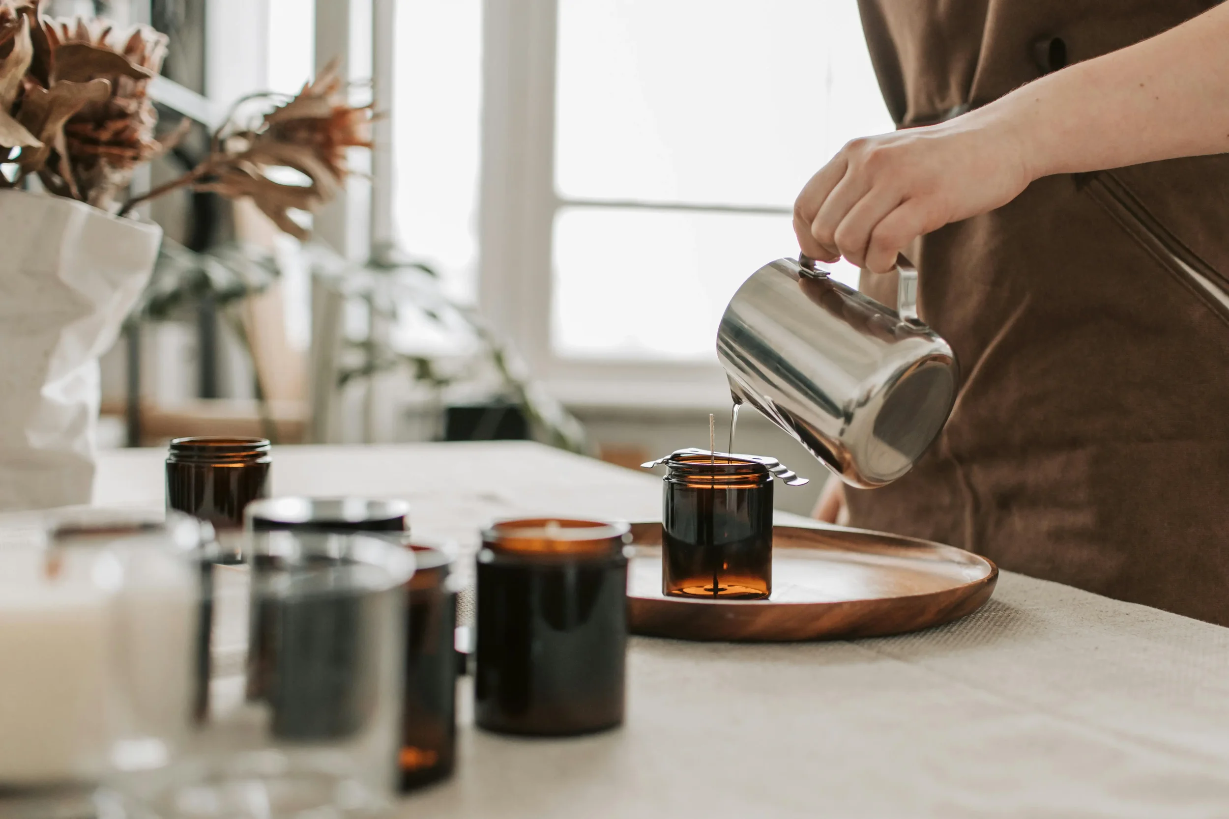 Person pouring liquid from a metal pitcher into a brown glass jar on a round wooden tray, with several other brown glass jars and containers on a beige tablecloth.