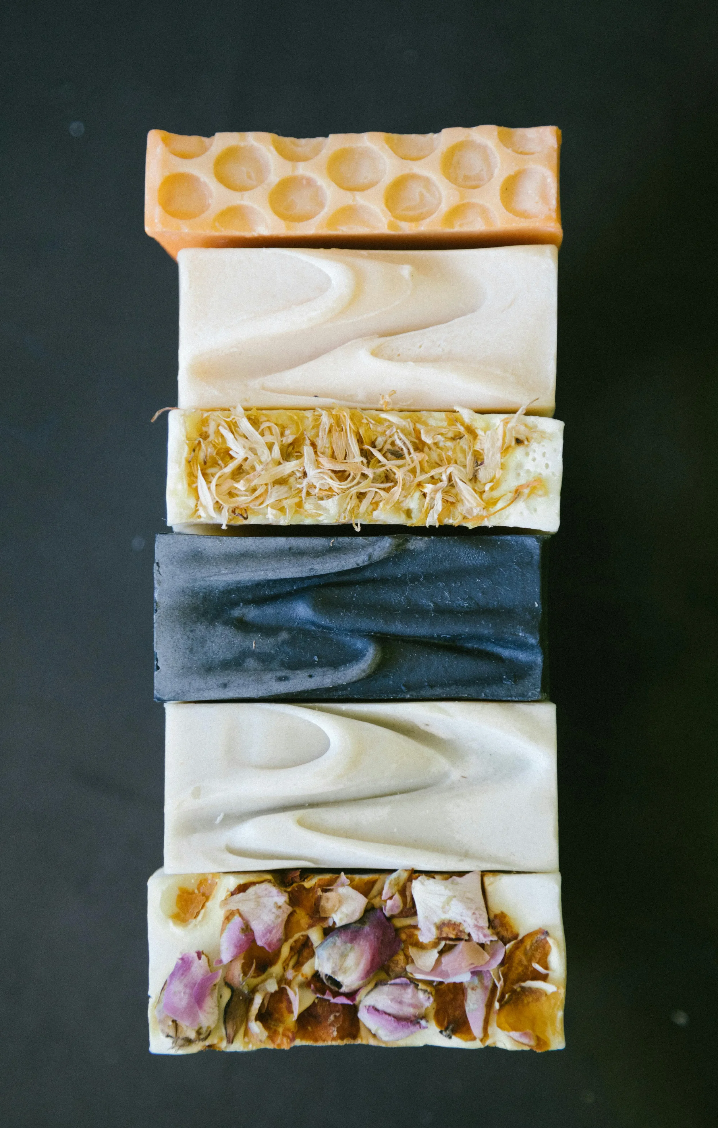 A vertical stack of six different handmade soap bars with various textures and colors, set against a dark background.