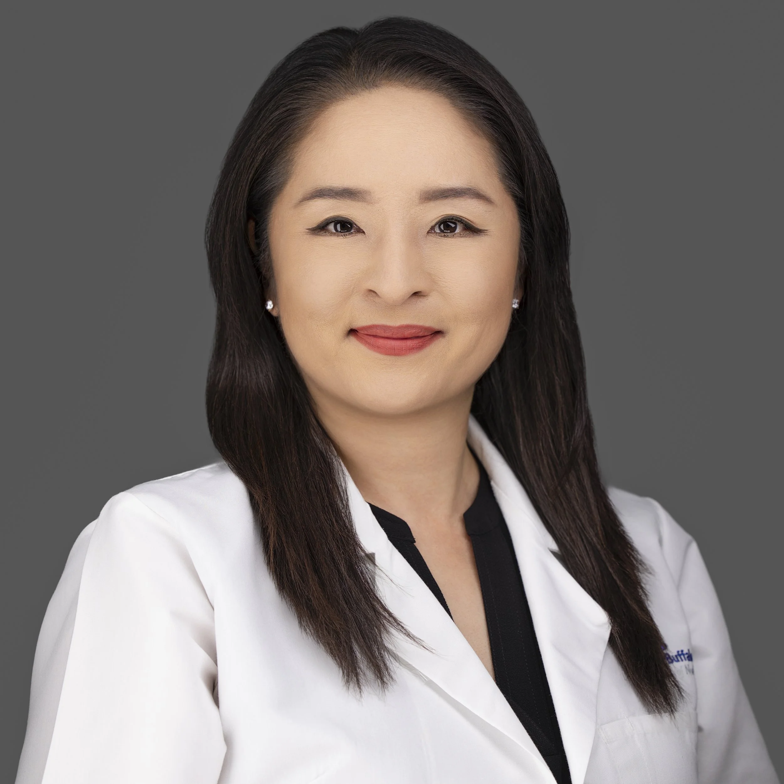 Professional woman with dark hair wearing a white coat, smiling against a gray background.