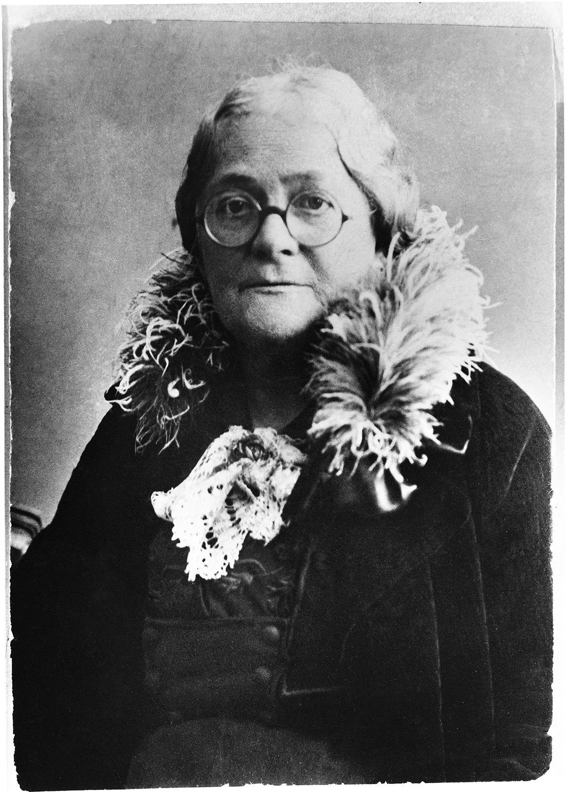 Black and white portrait of an older person with glasses, short hair, and a feathered scarf or collar, wearing a dark jacket.