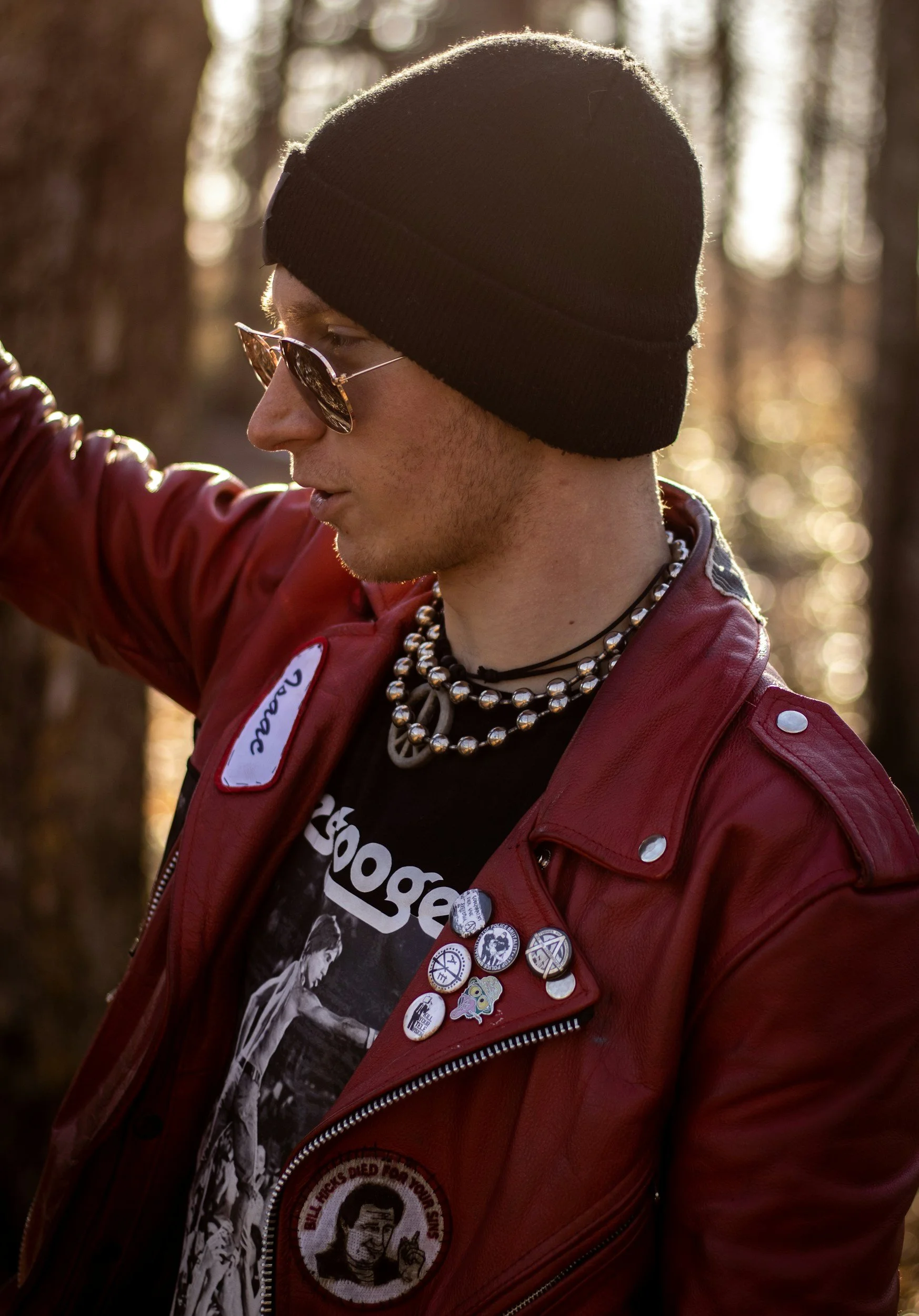 A young person wearing a black beanie, sunglasses, a red leather jacket with pins, layered necklaces, and a graphic T-shirt, standing outdoors with trees in the background.