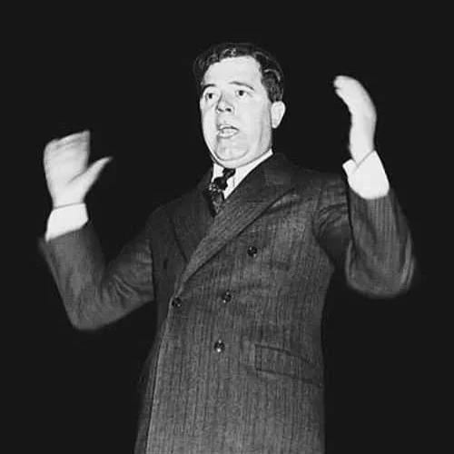 An elderly man with a weathered face and gray beard wearing a baseball cap and navy blue zip-up jacket outdoors, with wooden and brick background.