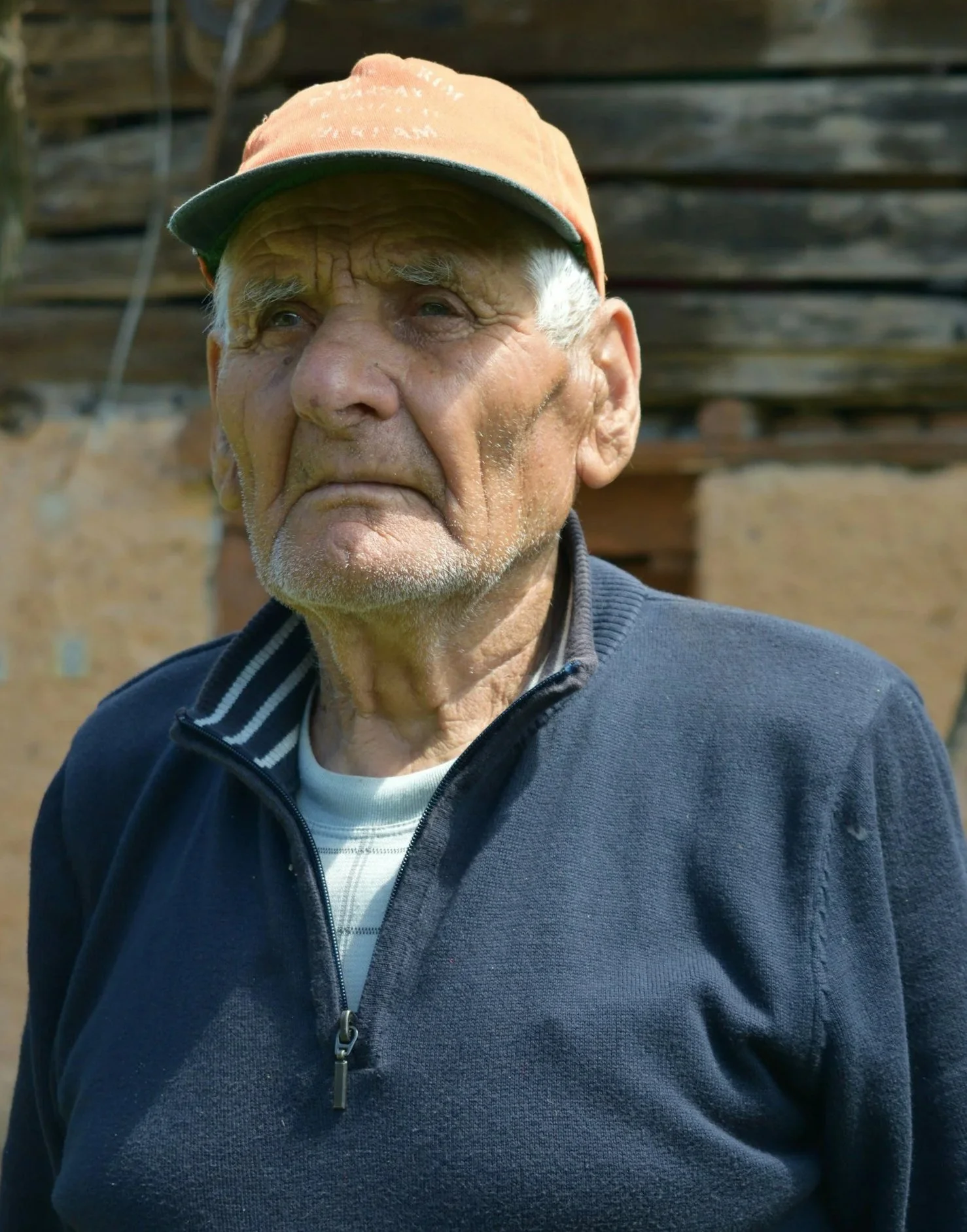 An elderly man with a weathered face and gray beard wearing a baseball cap and navy blue zip-up jacket outdoors, with wooden and brick background.