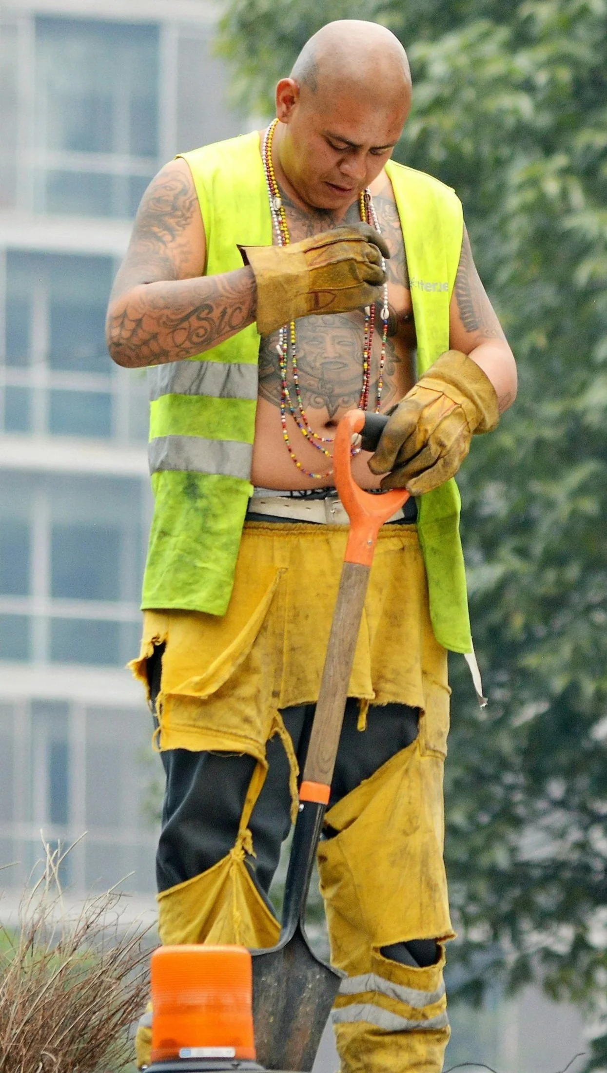 A construction worker standing outdoors, wearing a yellow safety vest and torn yellow work pants, is holding a trowel and looking down at it. The worker has tattoos on their chest and arms, and is wearing multiple necklaces.