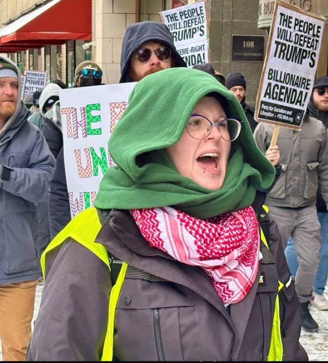 A person wearing glasses and a green hoodie protesting. She is surrounded by other protesters holding signs that say "The people will defeat Trump's billionaire agenda!". The person is also wearing a high-visibility vest.