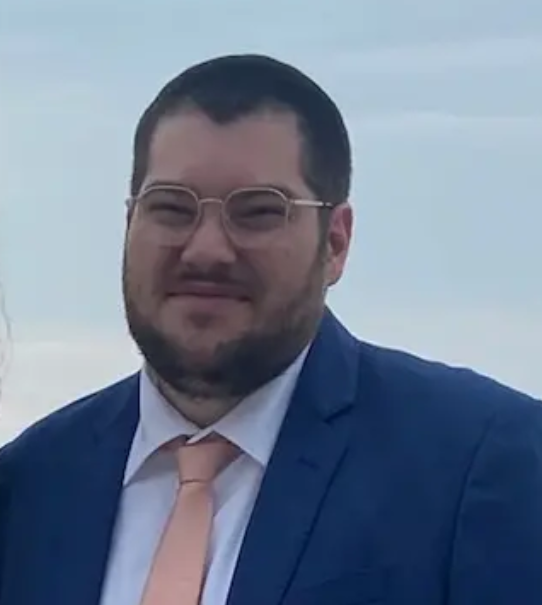 A construction worker standing outdoors, wearing a yellow safety vest and torn yellow work pants, is holding a trowel and looking down at it. The worker has tattoos on their chest and arms, and is wearing multiple necklaces.