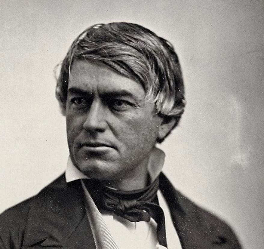 A young person wearing a black beanie, sunglasses, a red leather jacket with pins, layered necklaces, and a graphic T-shirt, standing outdoors with trees in the background.