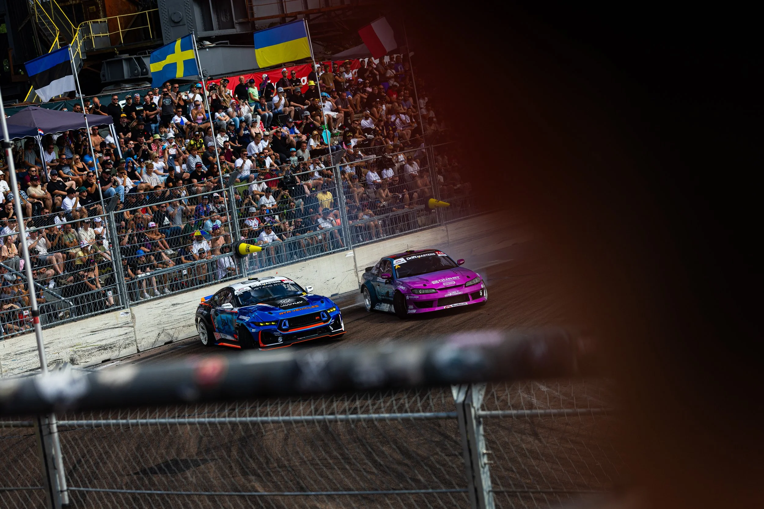 Two race cars, one blue and one purple, competing on a dirt track with a large crowd of spectators in the stands behind a chain-link fence, flags, and safety barriers.