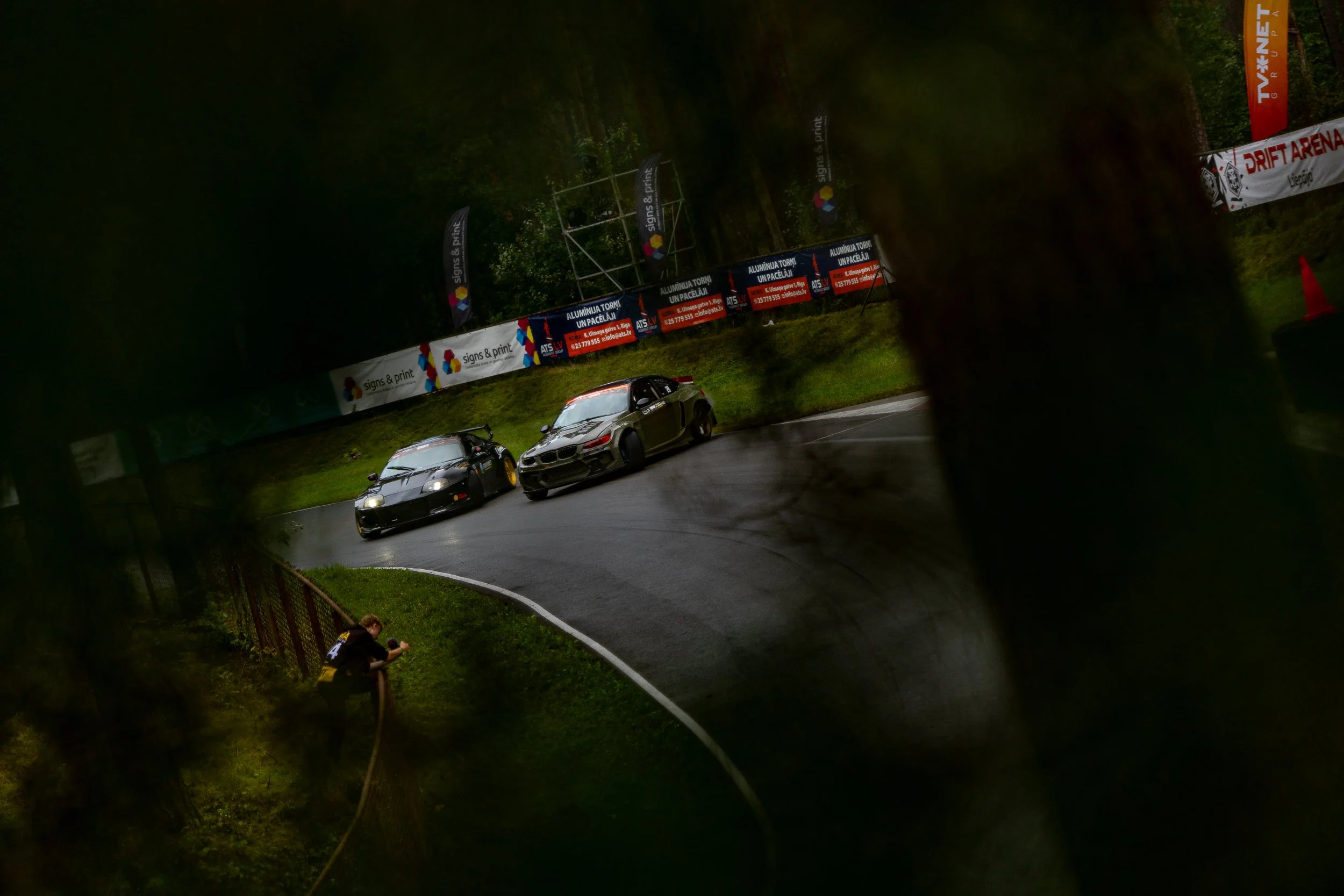 Two race cars on a wet, curved road track viewed through a gap between trees, with advertising banners in the background and a person with a camera near the fence.