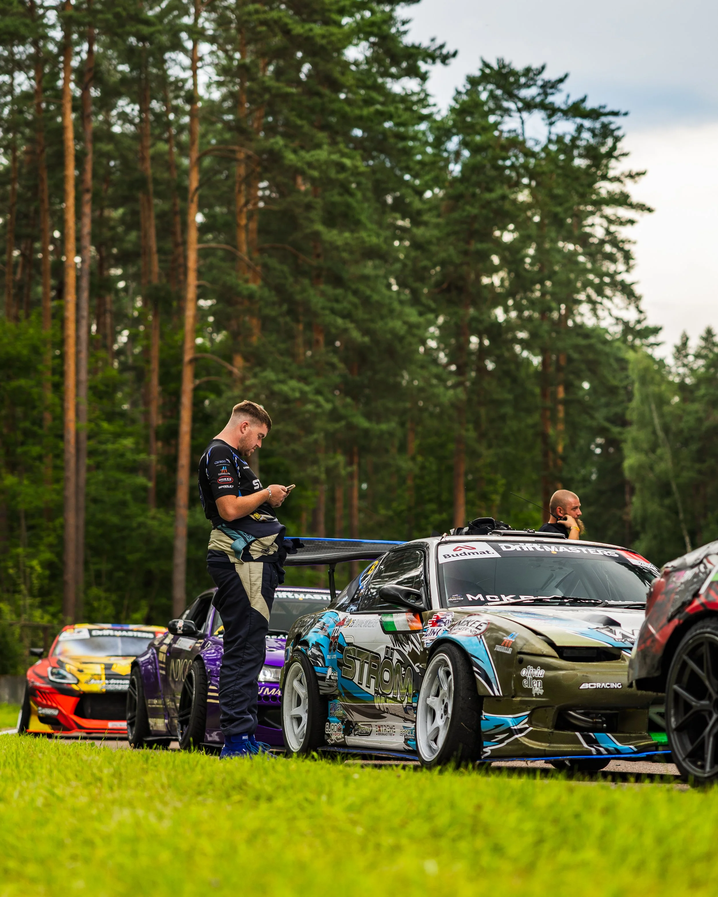 Two race car drivers preparing their cars on a grassy field, with a wooded forest in the background.