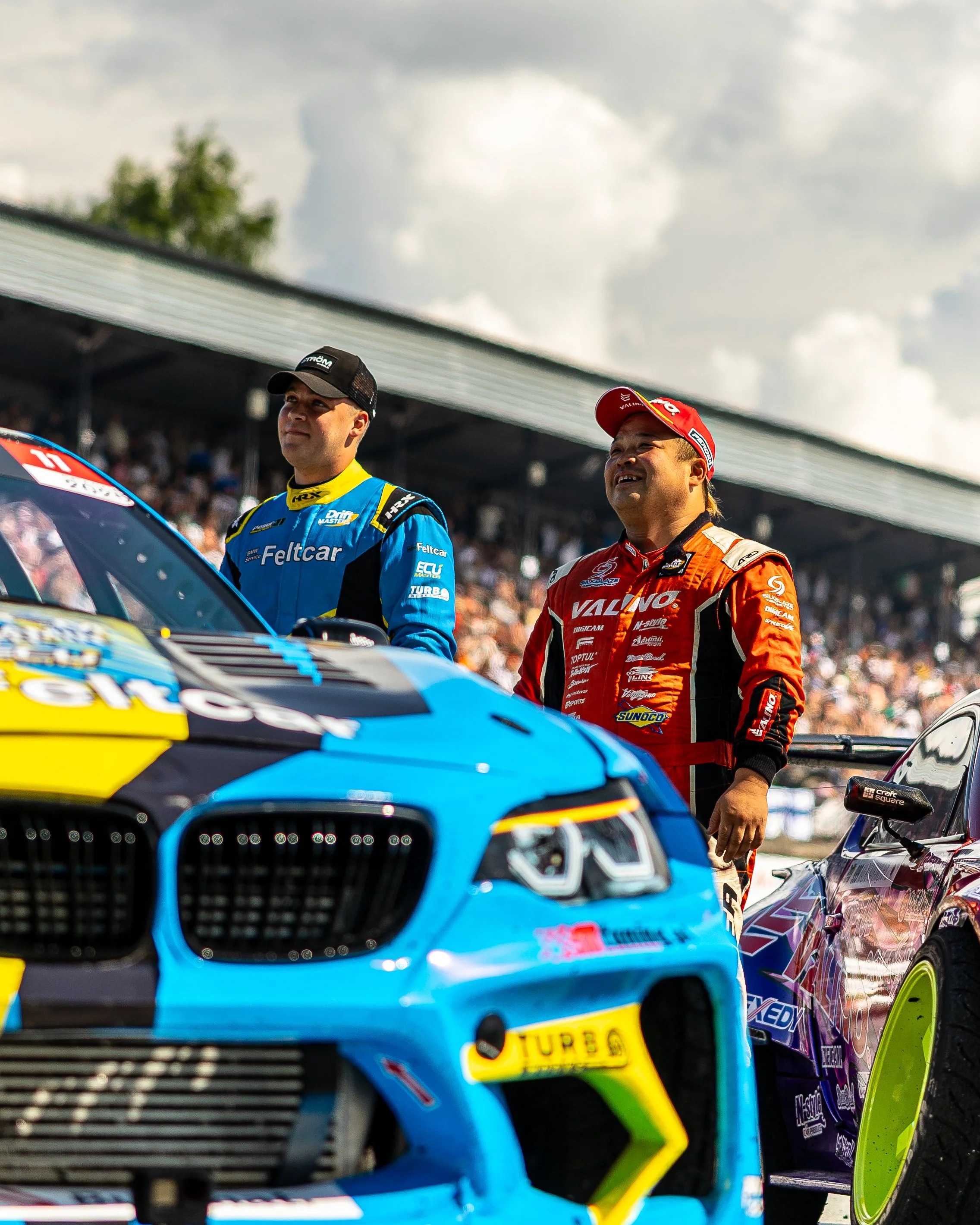 Two race car drivers standing next to their cars on a racing track, with a crowd in the background, one in a blue and yellow suit and the other in a red suit.