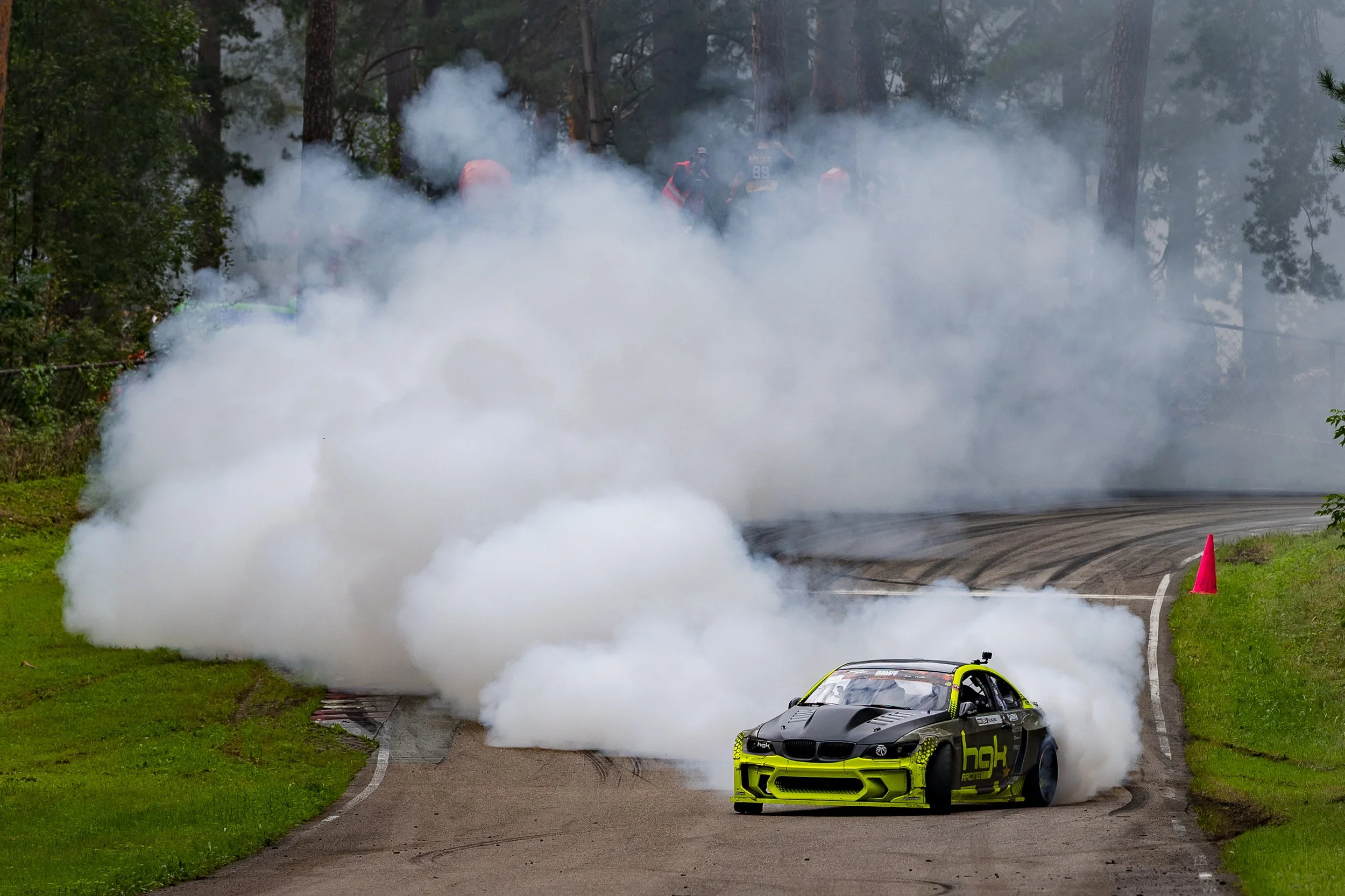 A race car drifts around a corner producing a large cloud of white smoke during a motorsport event, with safety cones marking the track boundary and a wooded area in the background.