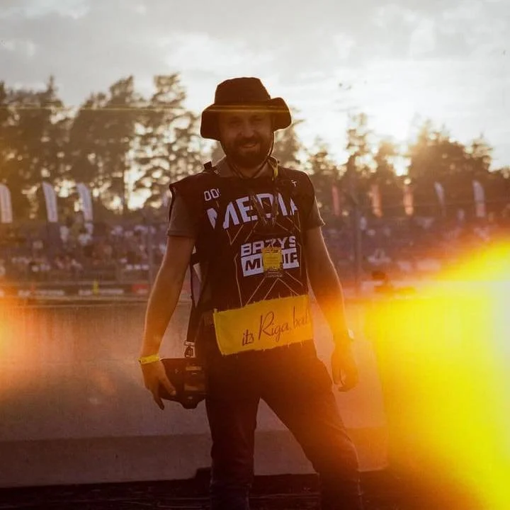 A man smiling and standing outdoors during sunset, wearing a black t-shirt with white text, a wide-brimmed hat, and a camera around neck, with a crowd and trees in the background and sunlight creating a lens flare effect.