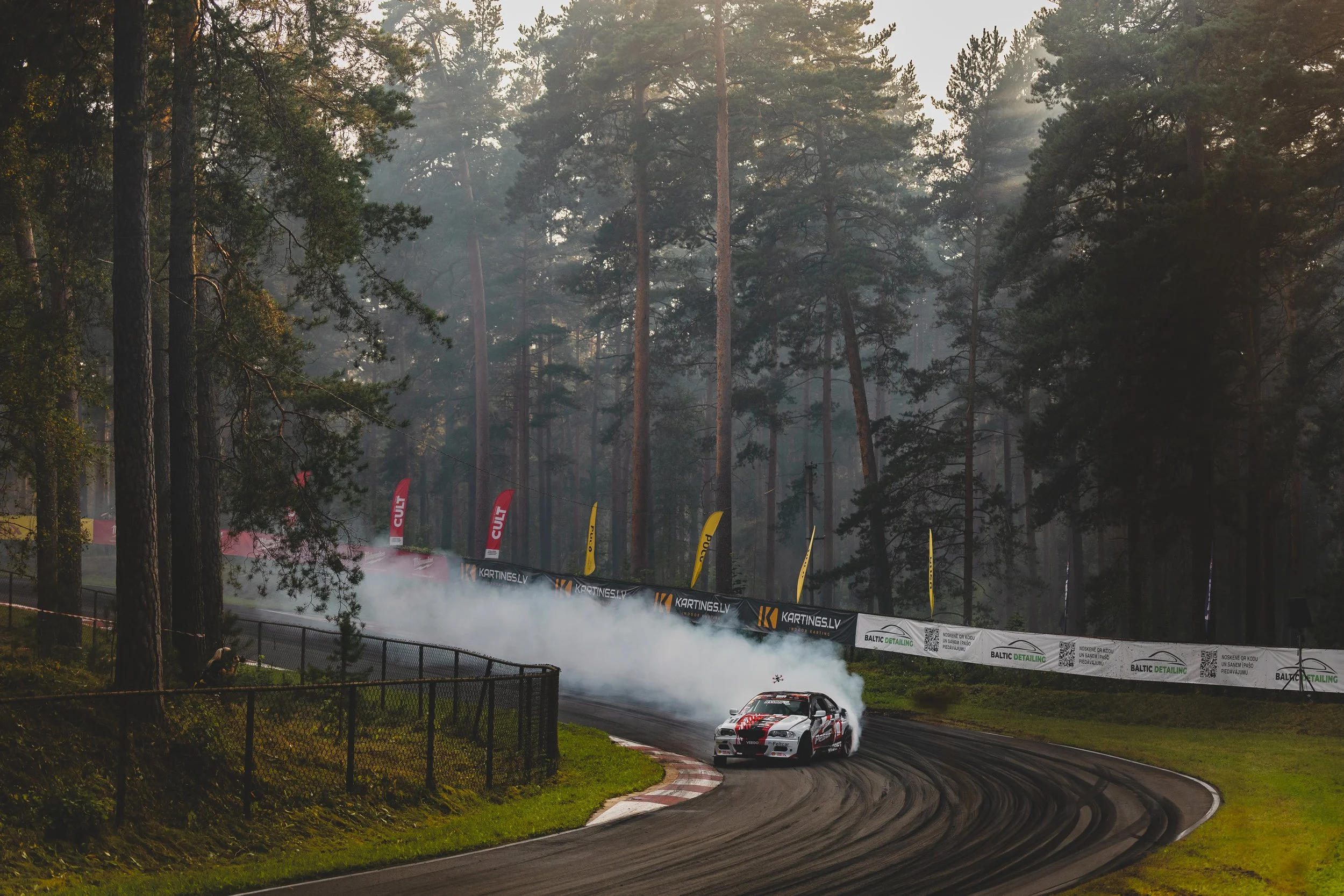 A race car drifting around a corner on a dirt track with tire smoke, surrounded by trees and banners along the fence.