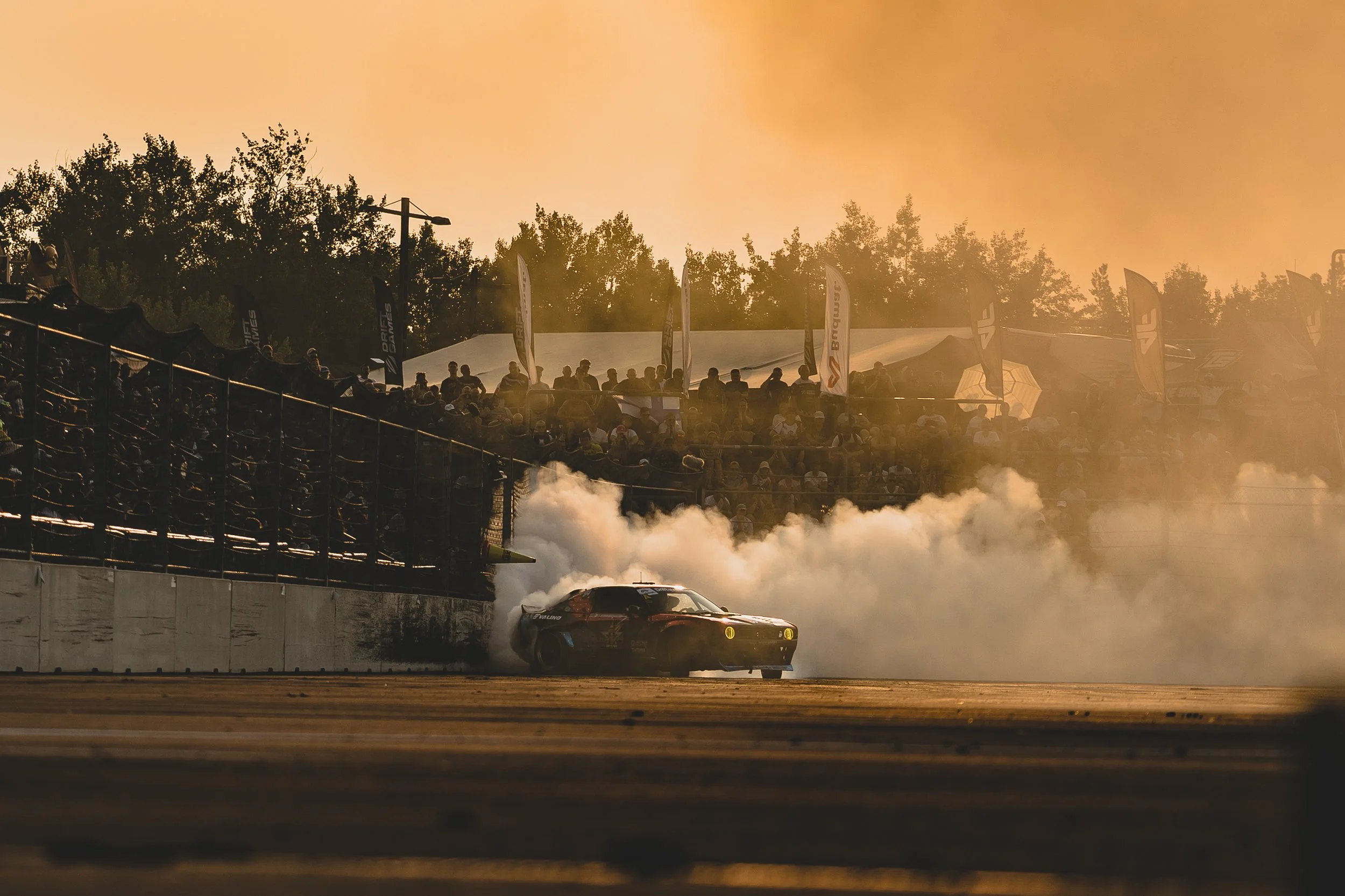 A race car drifting on a track during sunset, creating a cloud of smoke behind it while spectators watch from the grandstand.