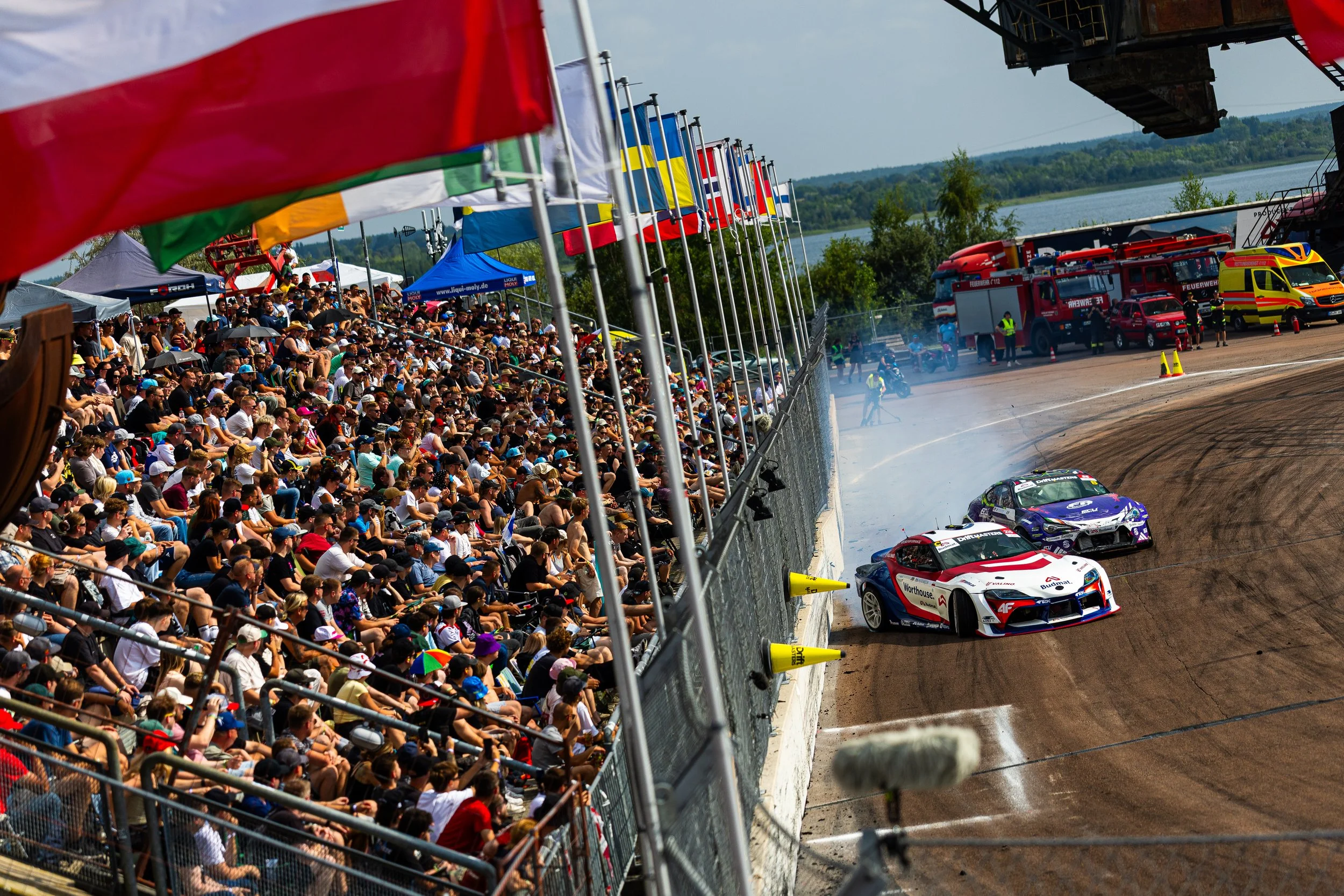Race cars on a dirt track during a motorsport event, with a large crowd of spectators watching from the grandstands.