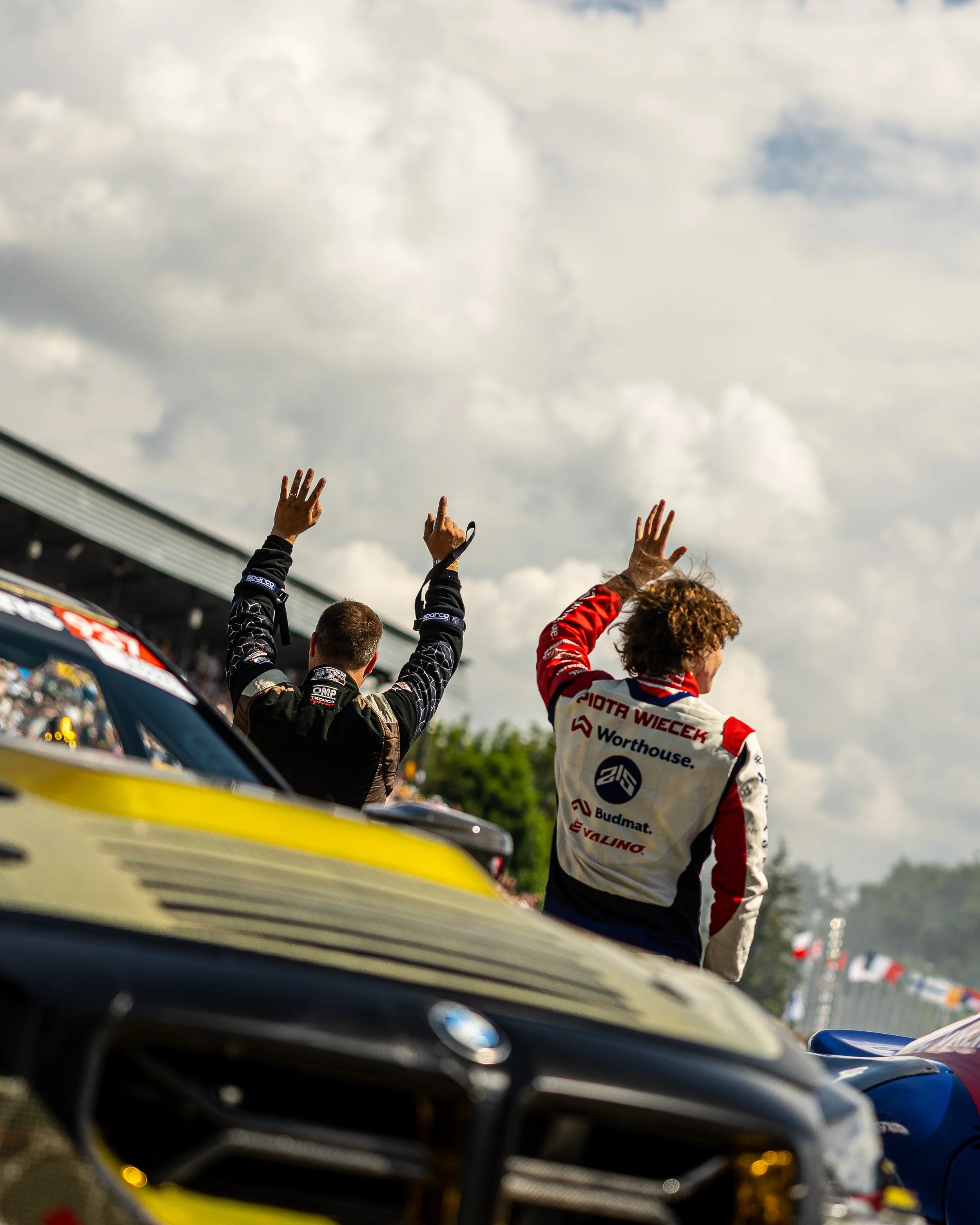 Two race car drivers standing on their cars, waving towards the crowd under a cloudy sky, during a motorsport event.