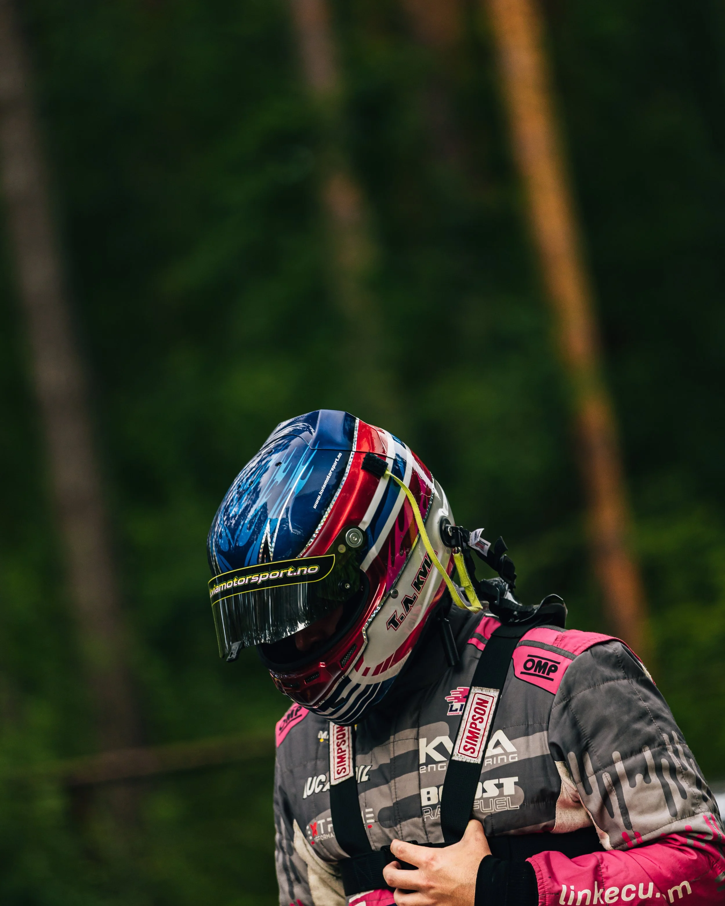 Motorsport racer in a pink and gray racing suit with a colorful helmet, standing in front of a blurred green forest background.