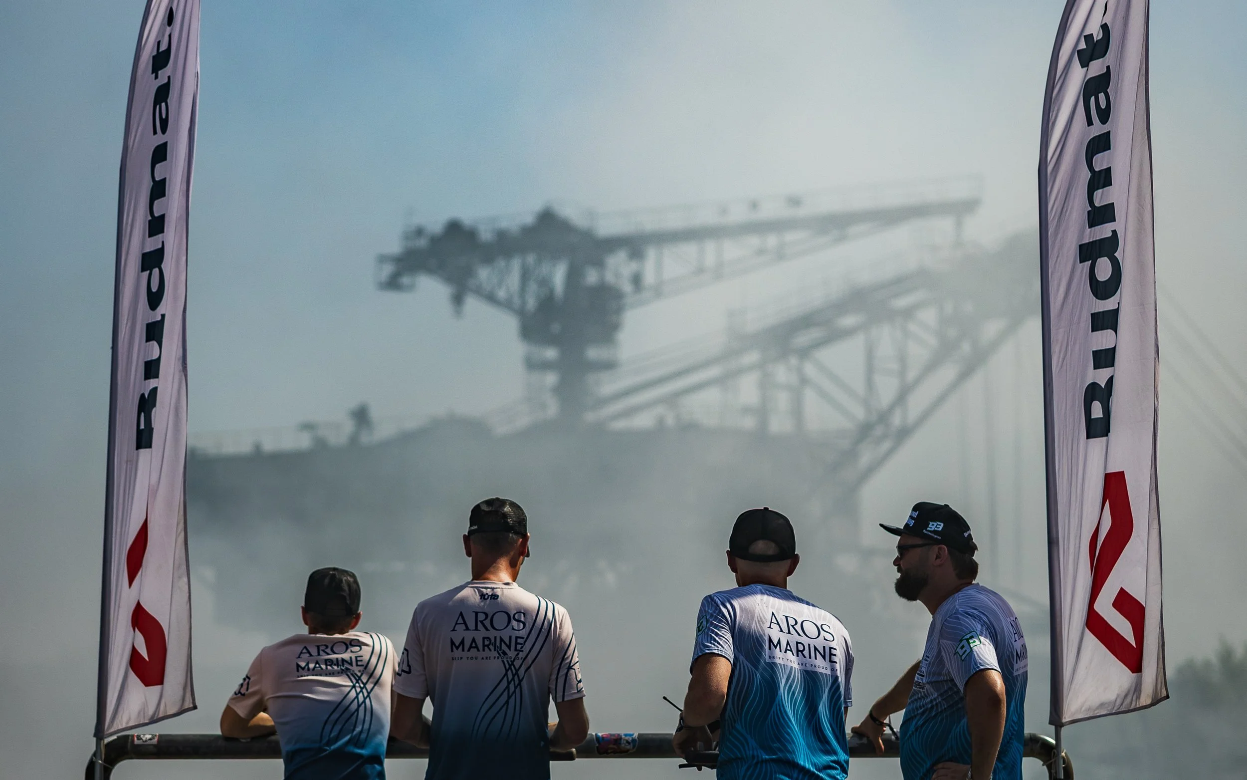 Four racing team members wearing Aros Marine shirts stand by a railing, watching a race or event, with large flags on either side bearing the BRM logo. In the background, a large industrial crane or mining equipment is shrouded in mist or smoke.