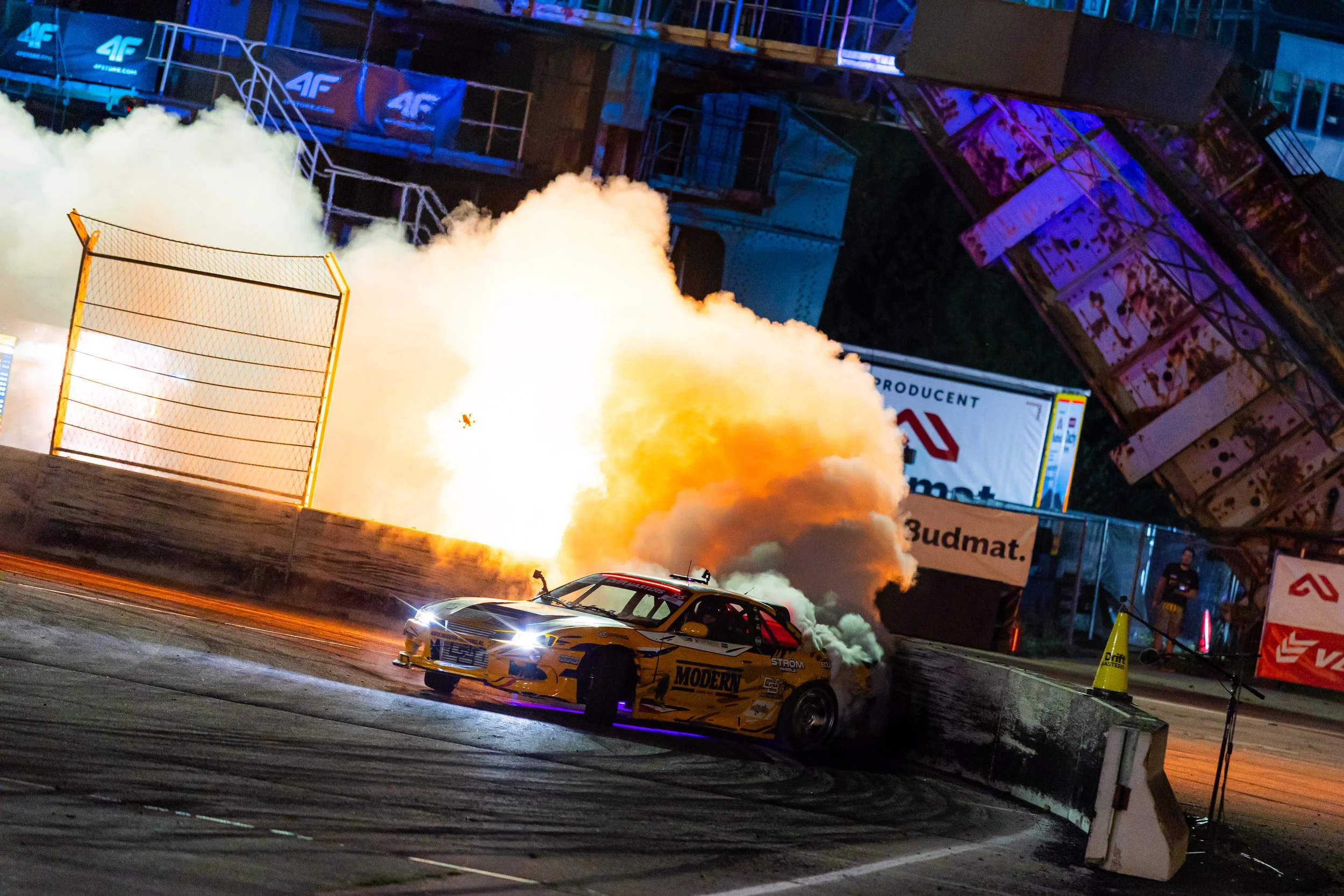 Race cars on a track, with one car emitting flames and smoke, at night with bright lights and barriers.