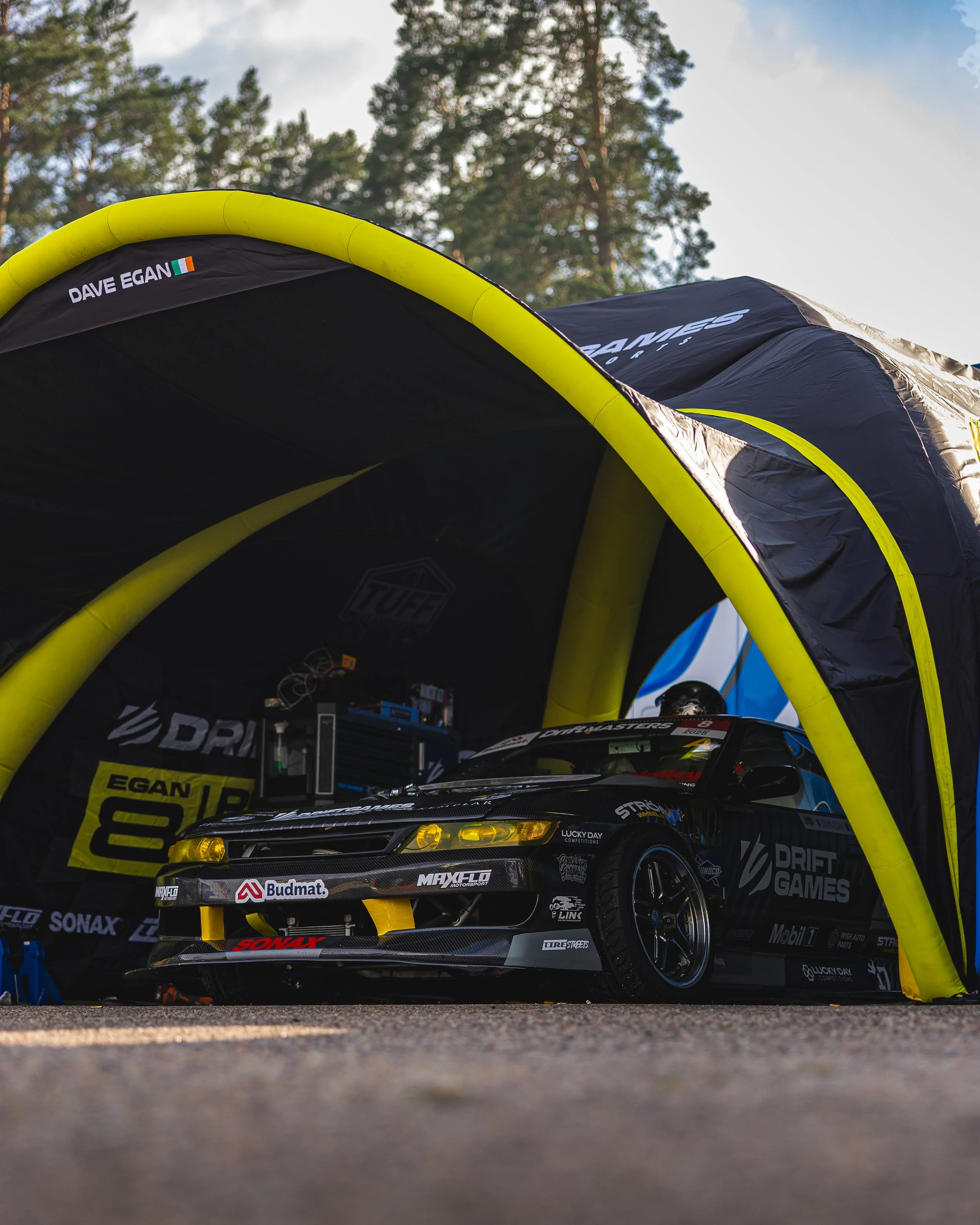 A race car under a yellow and black tent, with various sponsor logos, in a wooded outdoor area.