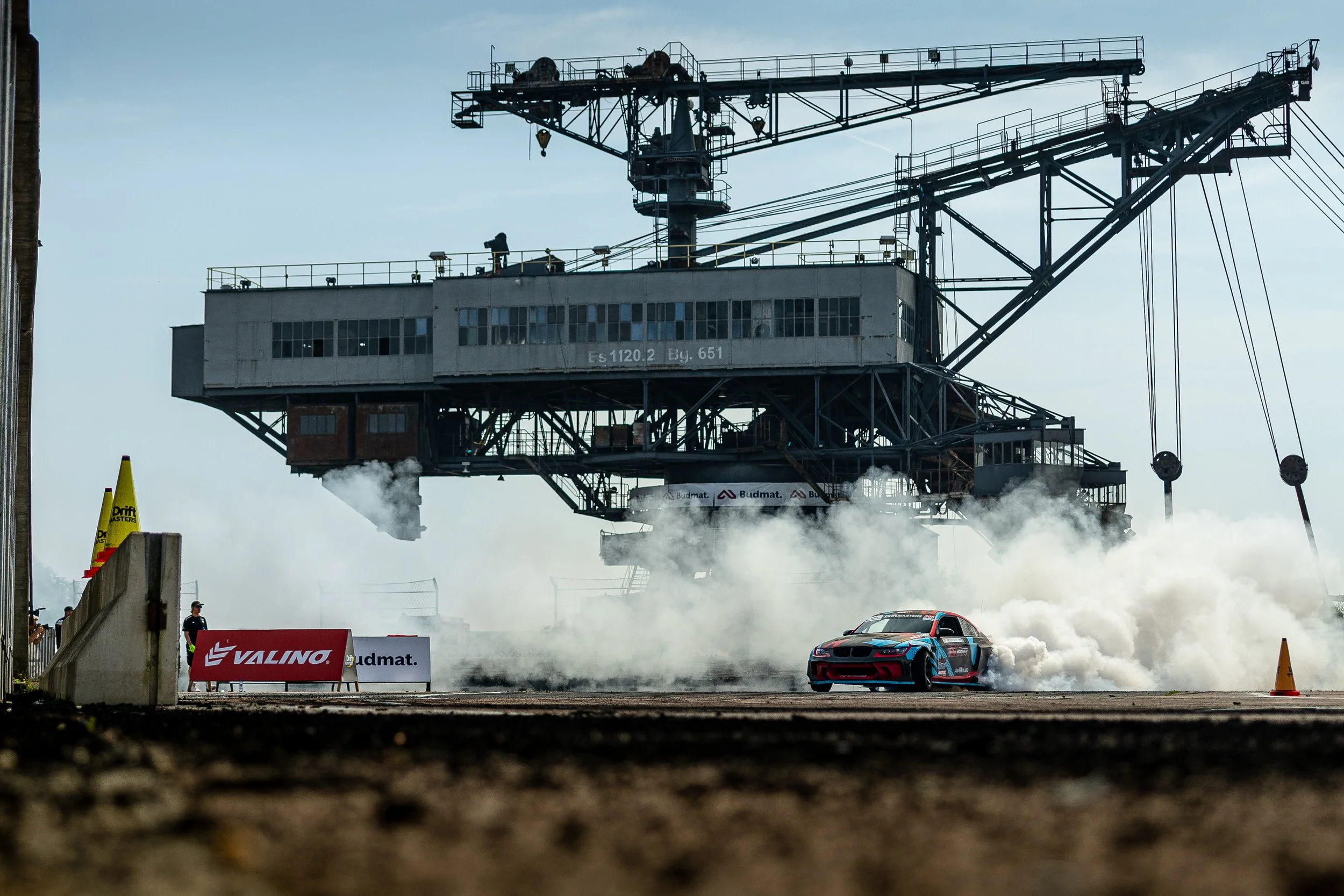 Race car drifting on a track with smoke, large industrial crane overhead, and event banners visible.