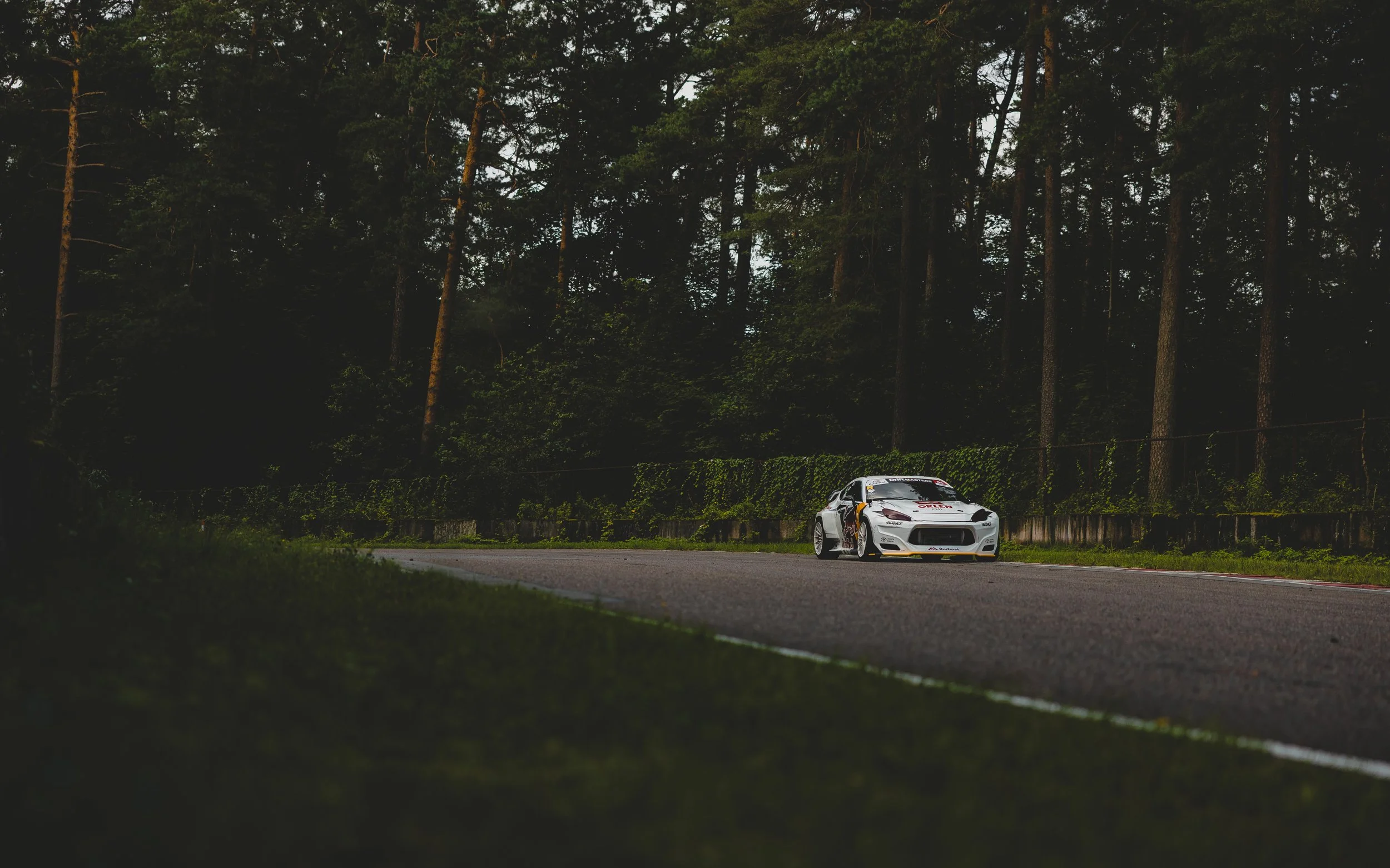 A white race car on a track surrounded by tall trees and greenery
