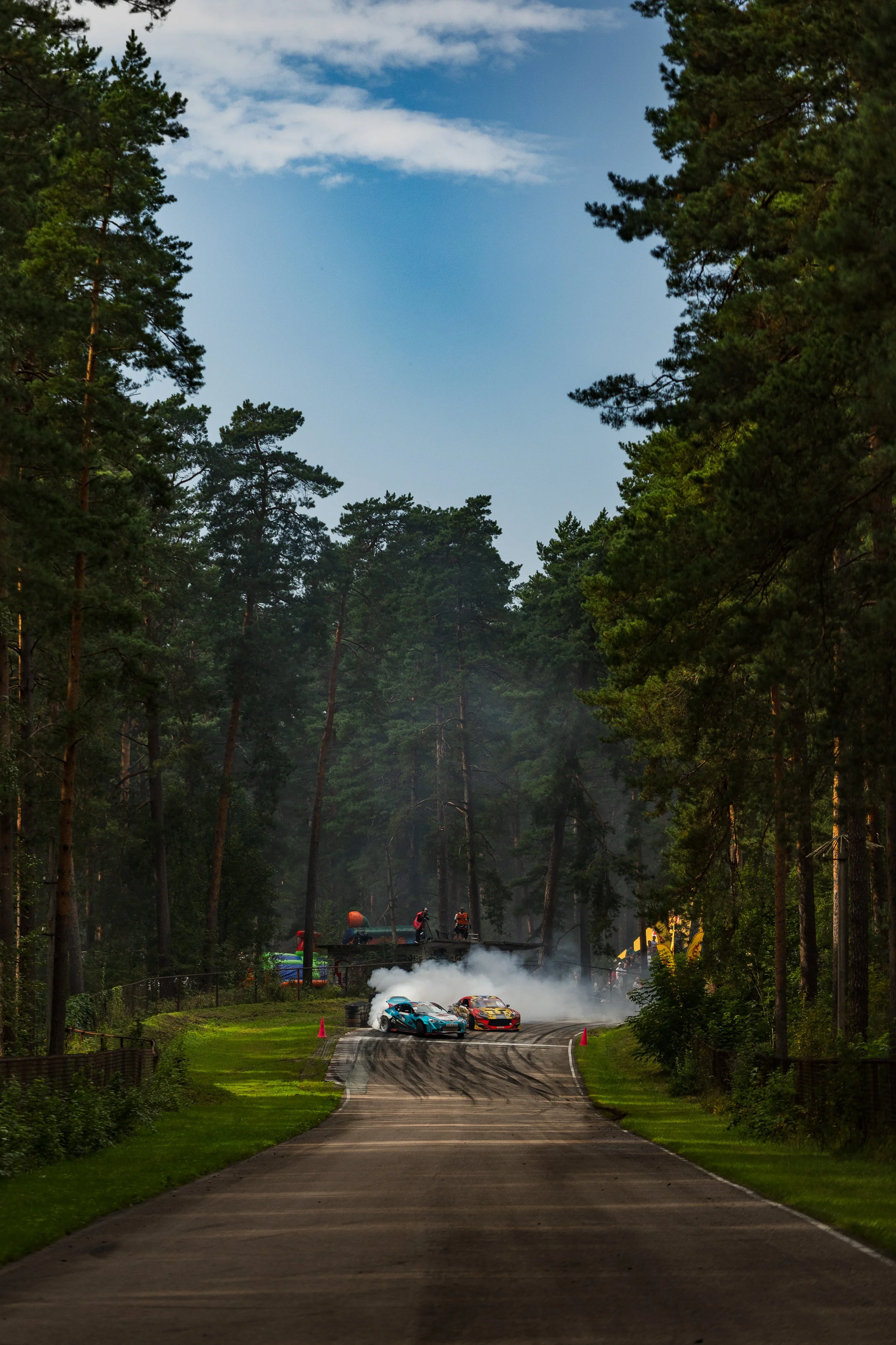 Two race cars drifting around a curve on a forested race track with tire smoke and spectators in the background.