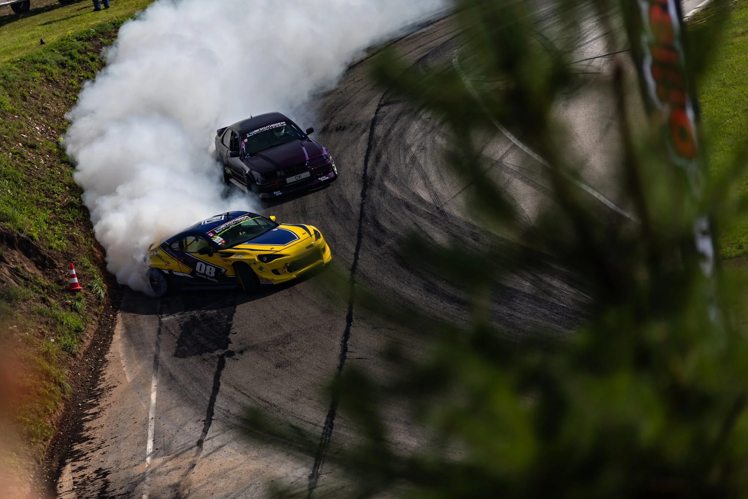 A yellow and blue race car drifting on a dirt racetrack, emitting smoke from the tires, with a black car nearby and blurred green leaves in the foreground.