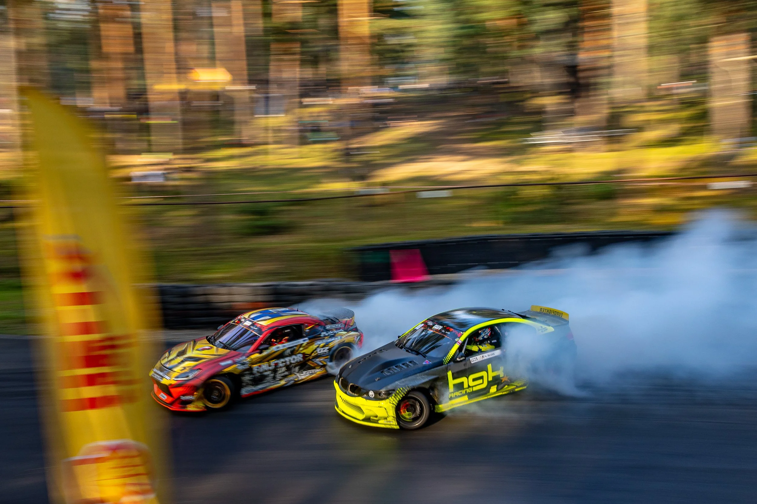 Two race cars competing on a track, with one car drifting and creating smoke, during a motorsport event.