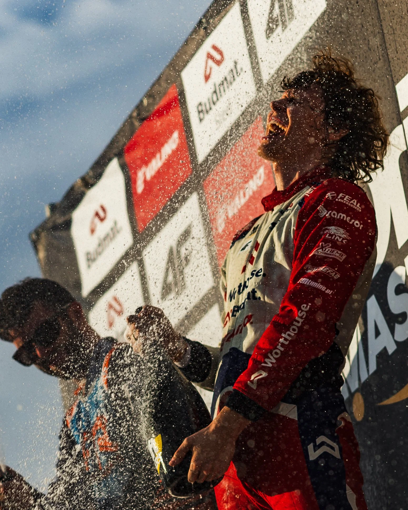 Two race car drivers celebrating on the podium with champagne, with droplets flying and a sponsor banner in the background.
